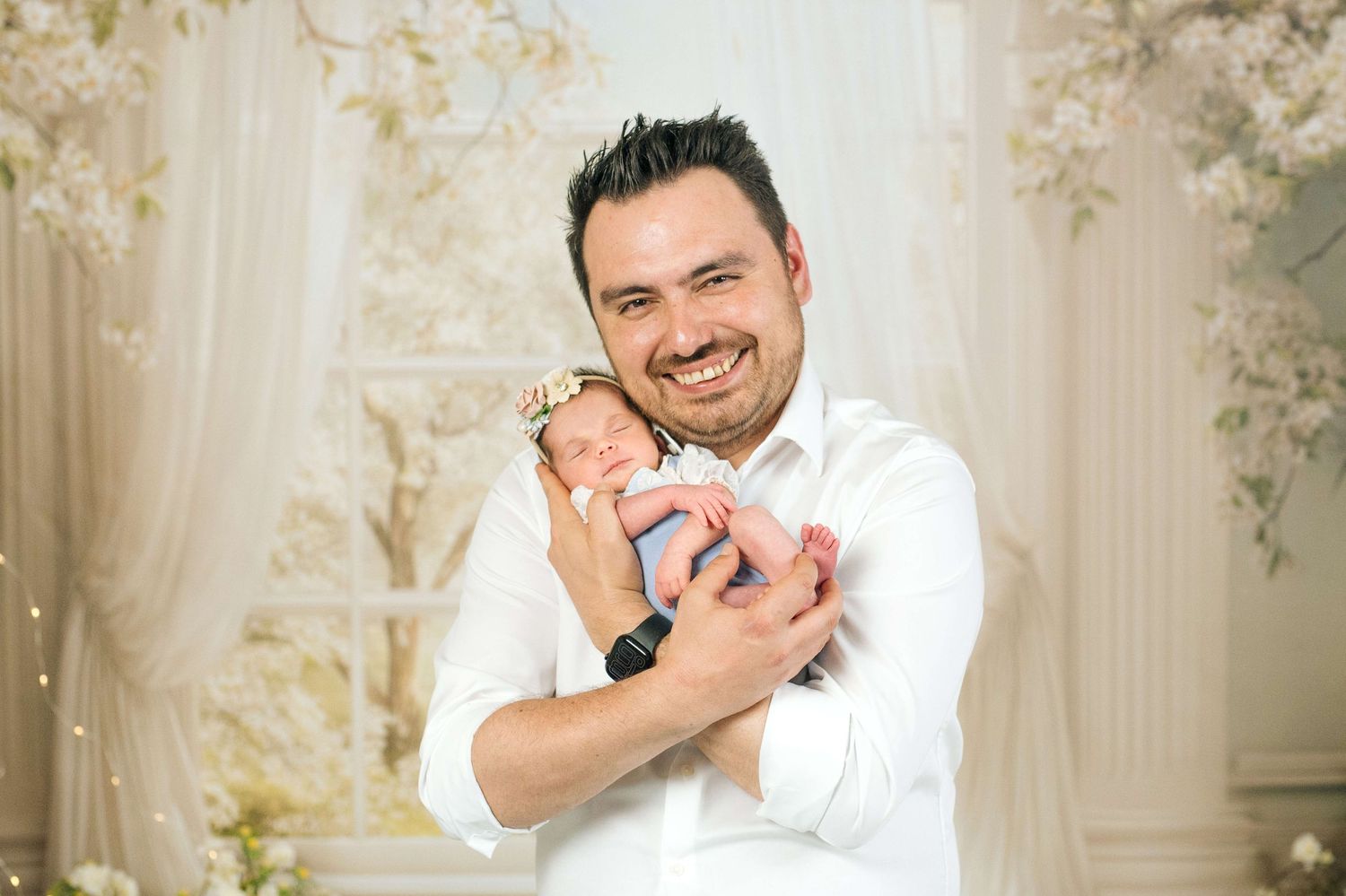 Smiling man in white shirt proudly holds sleeping newborn baby during a professional studio photo session.