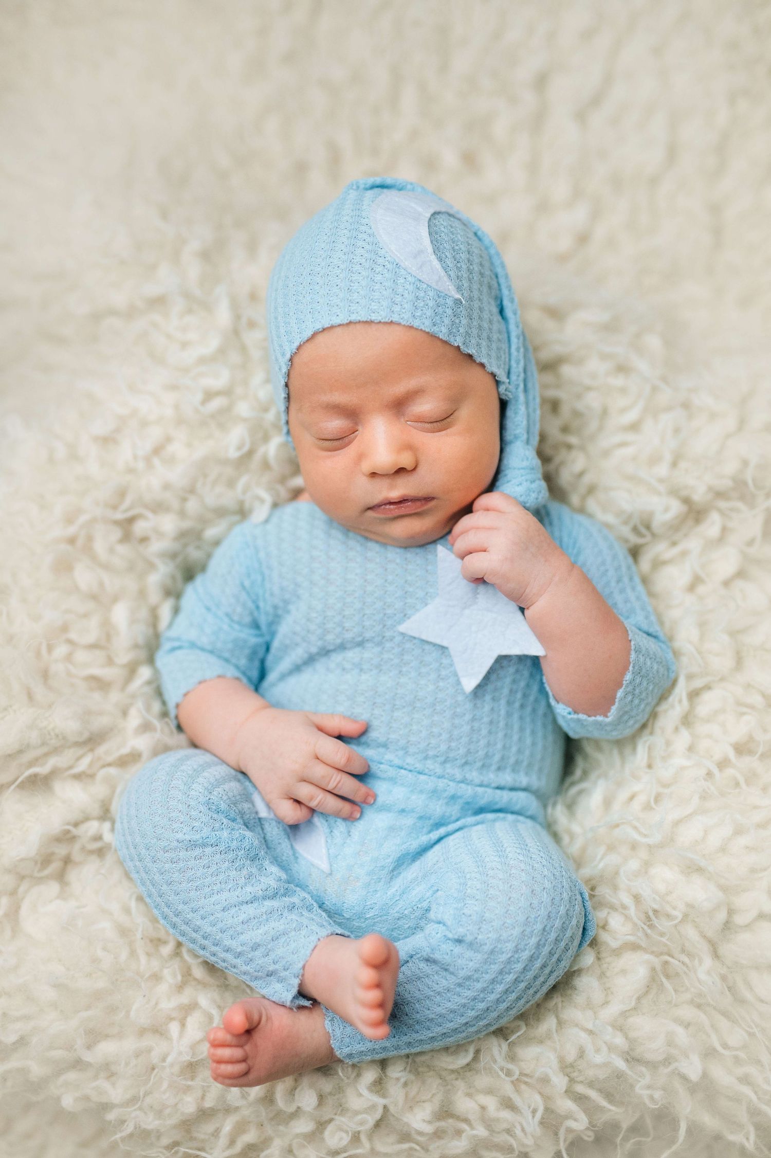 Newborn baby sleeping peacefully in blue knit outfit and hat, holding a star on a fluffy cream blanket.