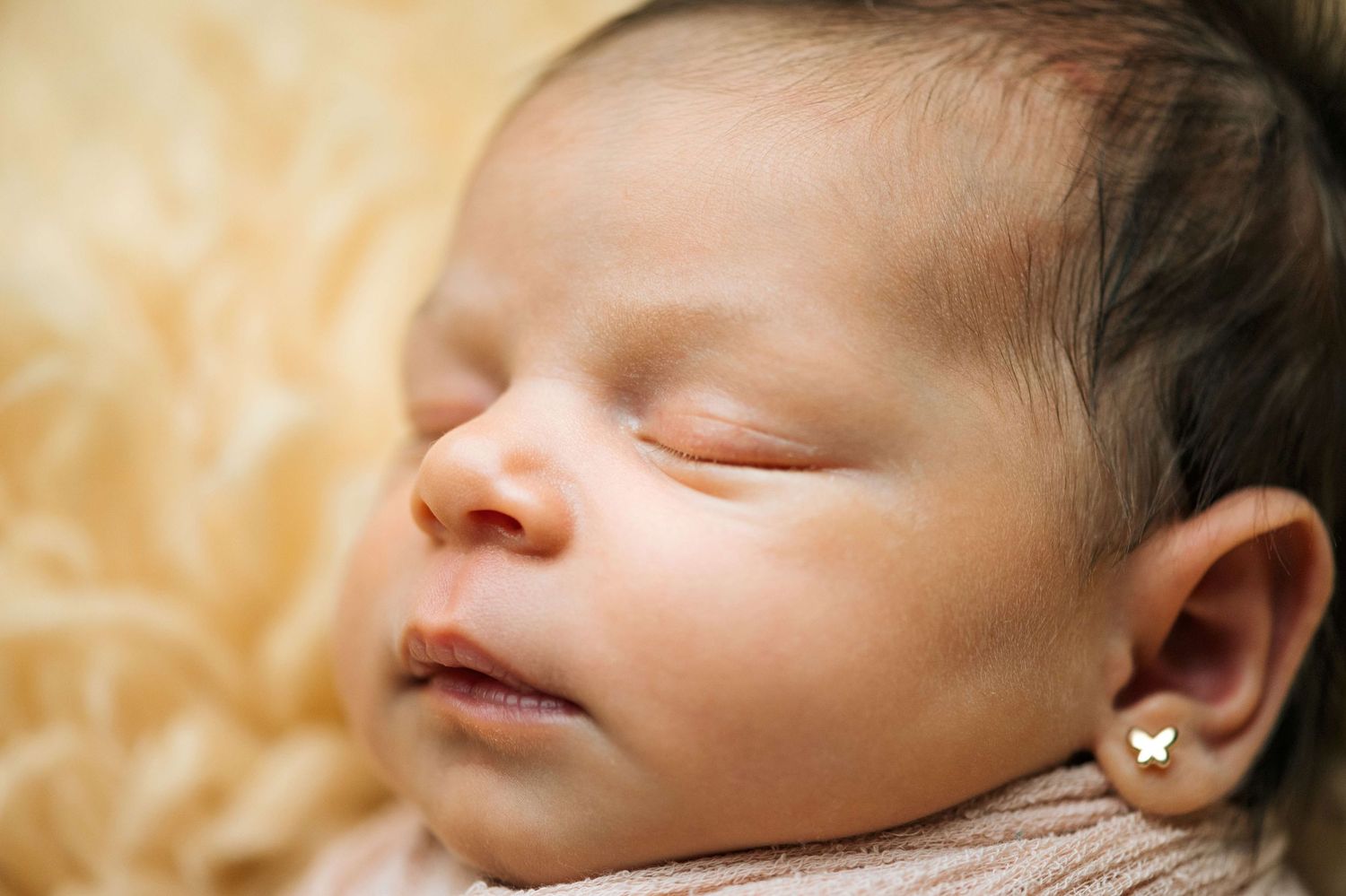 Peaceful sleeping newborn baby with tiny butterfly earrings resting on a soft fluffy cream blanket.