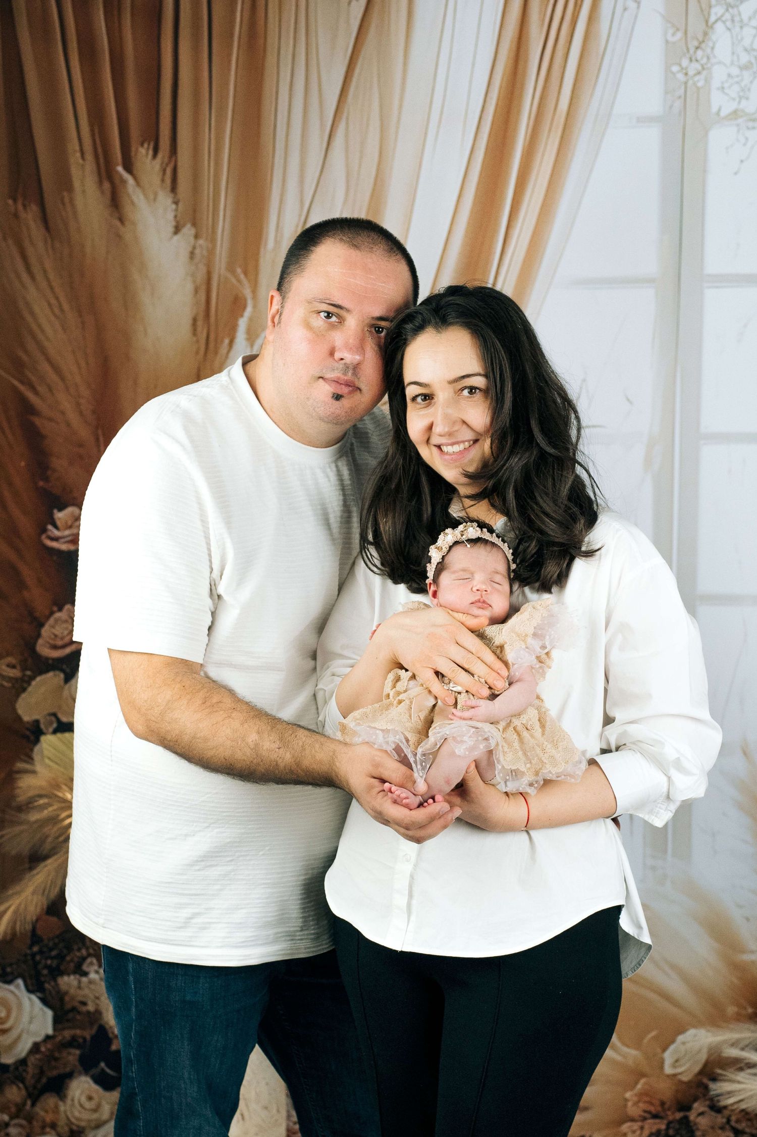 Happy parents in white shirts hold their sleeping newborn baby girl in a lace outfit during a studio family photo session.