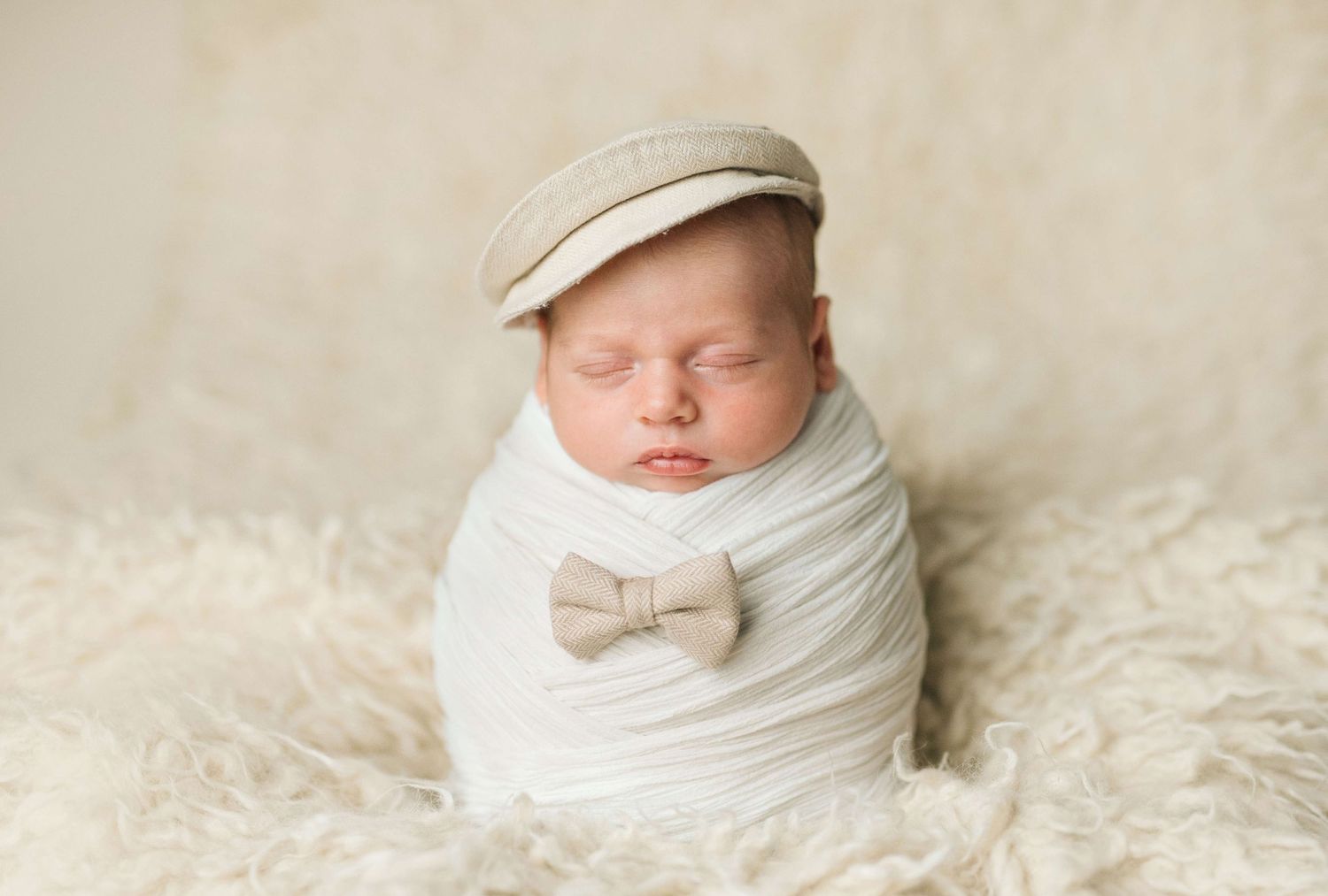 Sleeping newborn baby wrapped in white swaddle wearing a beige flat cap and bow tie on a fluffy cream rug.