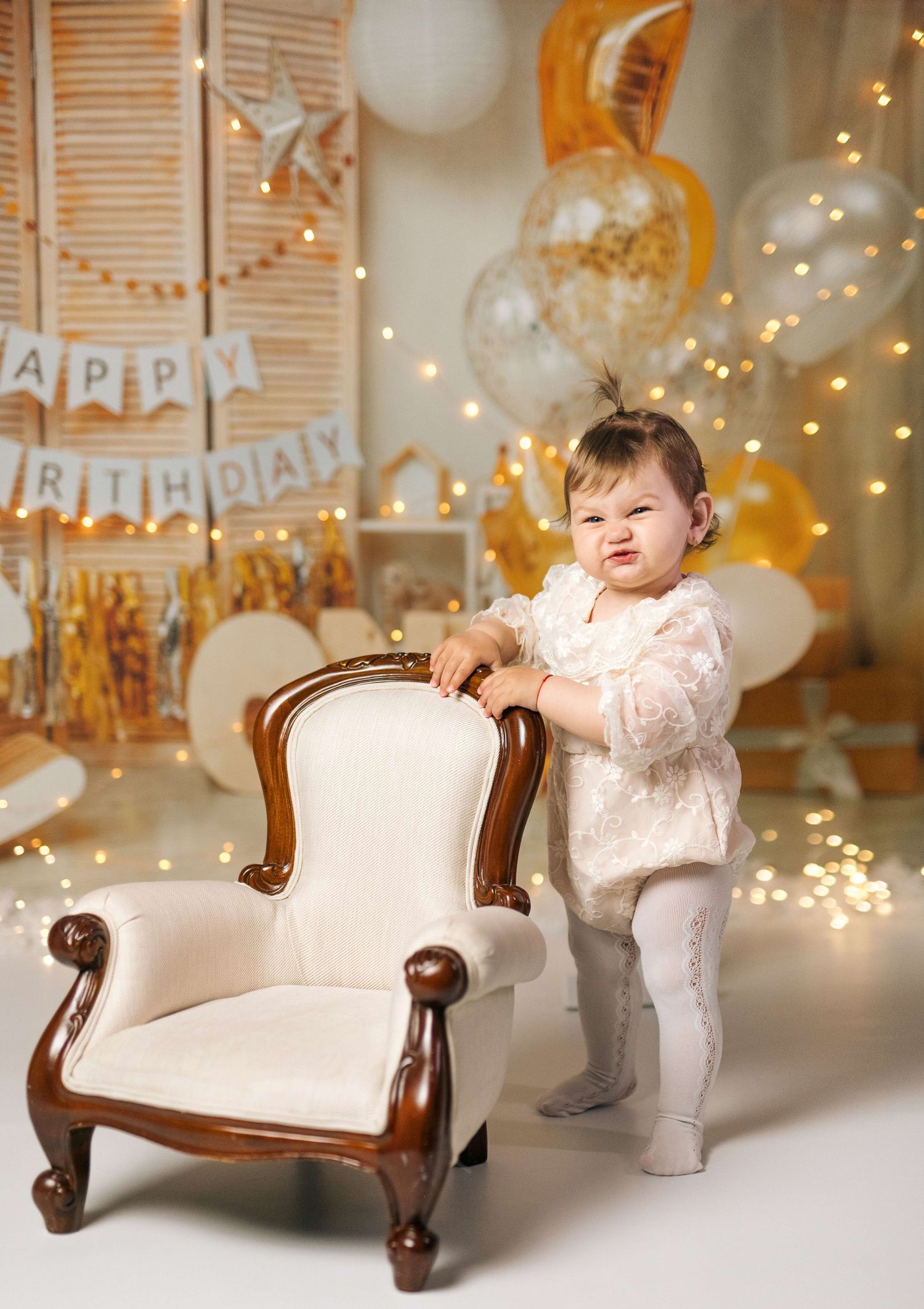 A baby girl in a white outfit stands by an elegant chair at a first birthday party with gold balloons and Happy Birthday banner.