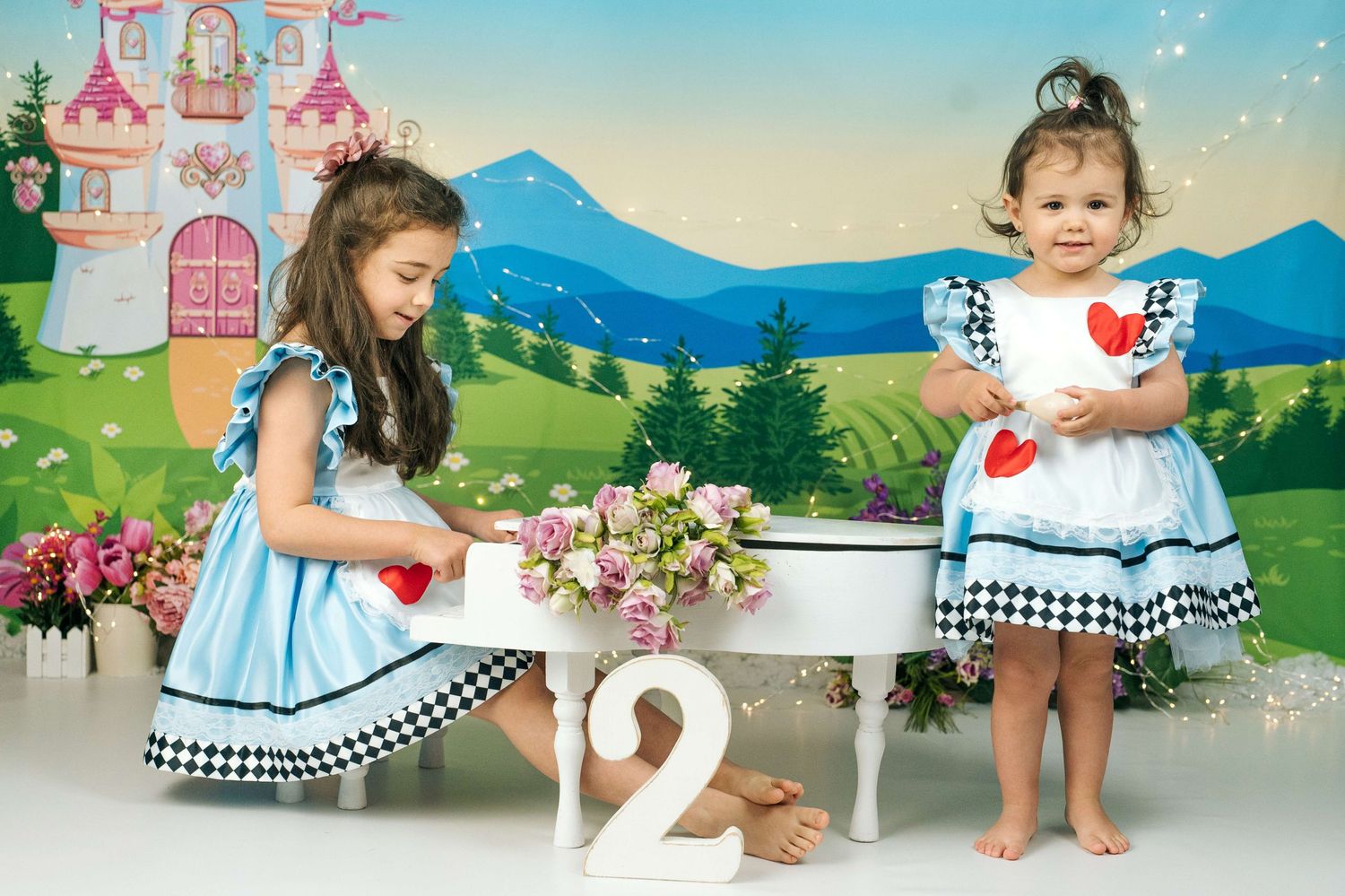 Two little girls in matching Alice in Wonderland outfits stand by a white piano with number 2 decoration.