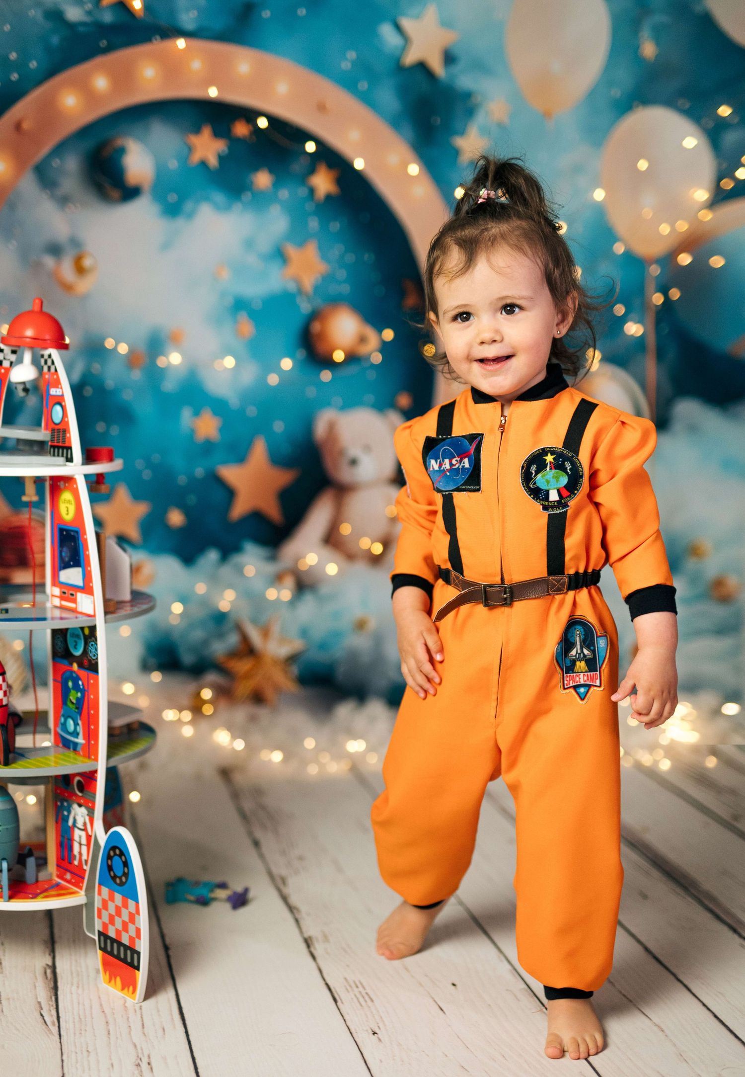 Toddler girl in orange NASA astronaut costume smiling in front of a space-themed backdrop with stars and rockets.