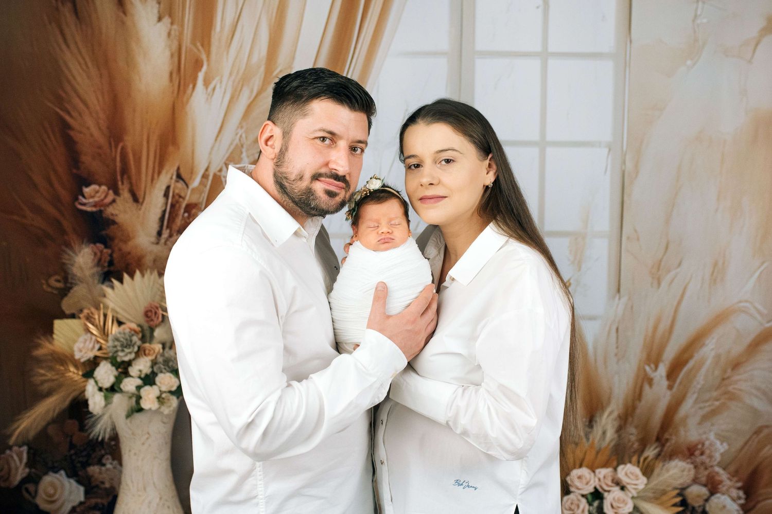 Happy couple in white outfits holding their newborn baby in a boho-style photo studio with floral decor.