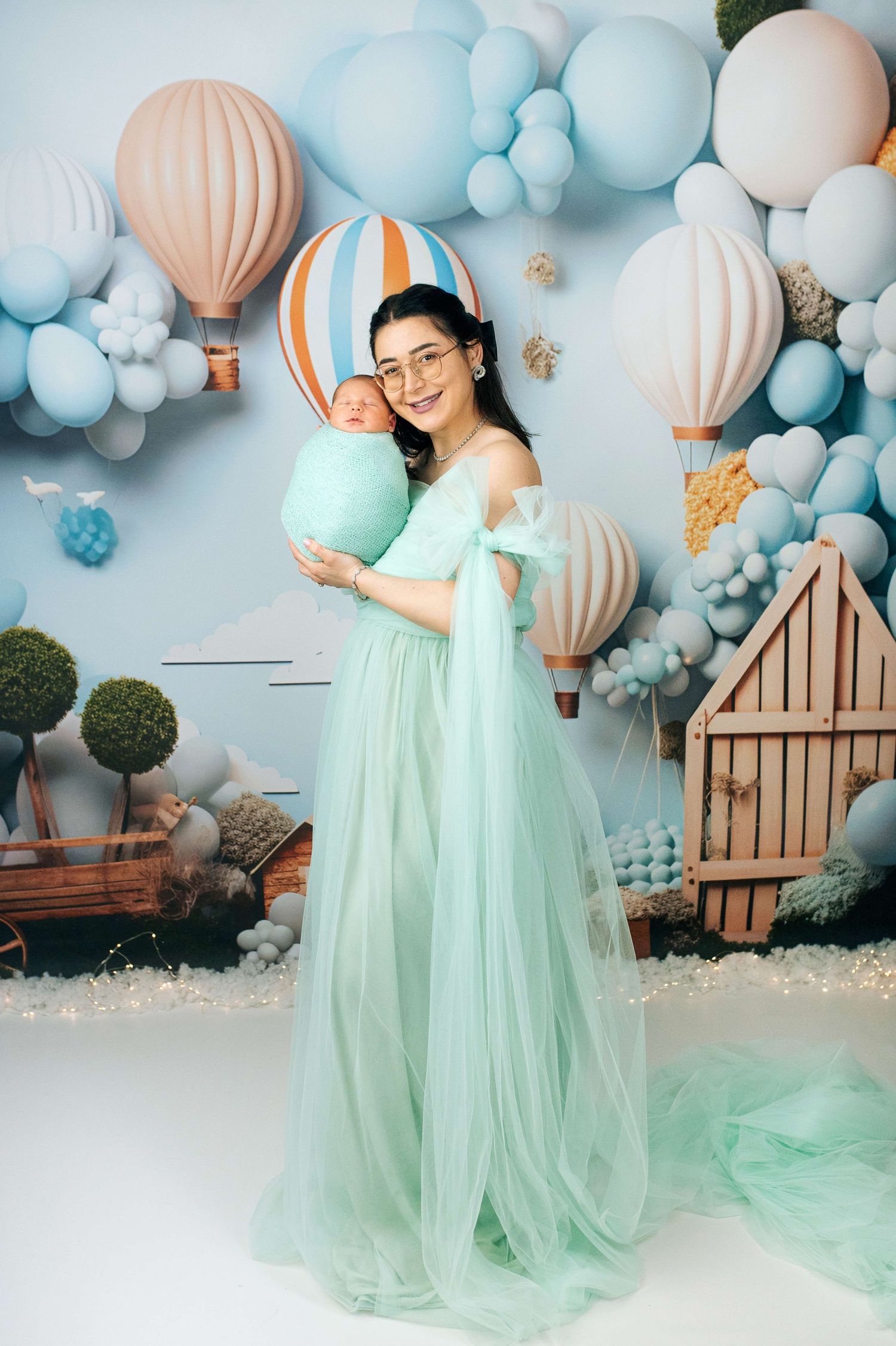 A mother in a mint green tulle gown holds her newborn baby amid a whimsical hot air balloon backdrop.