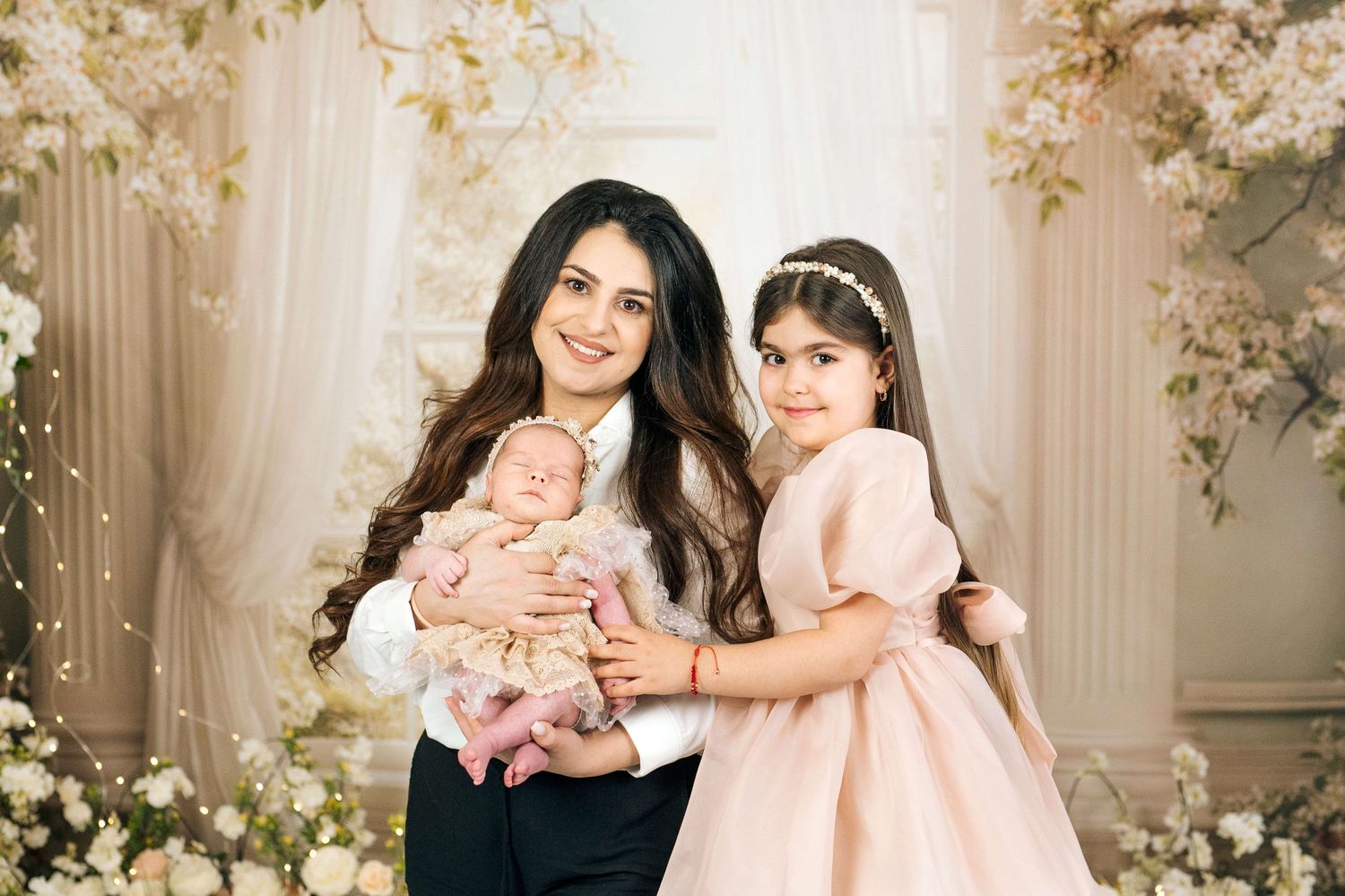 A woman in black and white outfit holds a newborn baby beside a young girl in pink dress, posing in floral studio setting.
