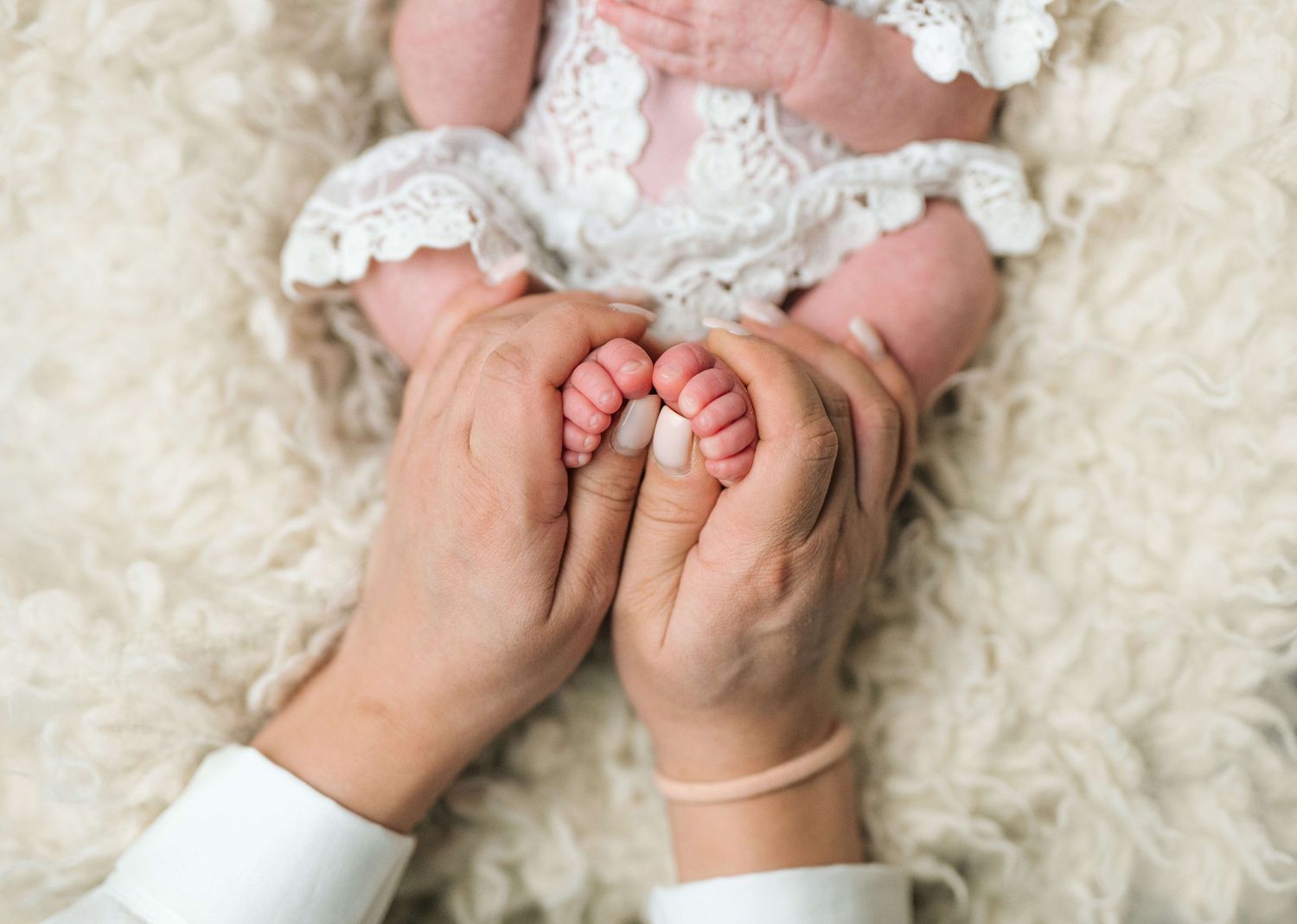 Adult hands gently cradling tiny newborn baby feet in a soft white lace outfit on a fluffy cream blanket.