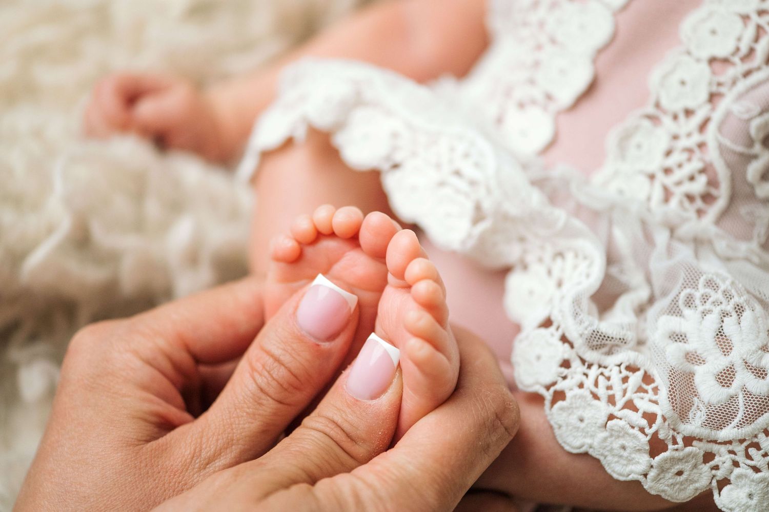 Close-up of adult hands gently holding tiny newborn baby feet in a white lace dress.