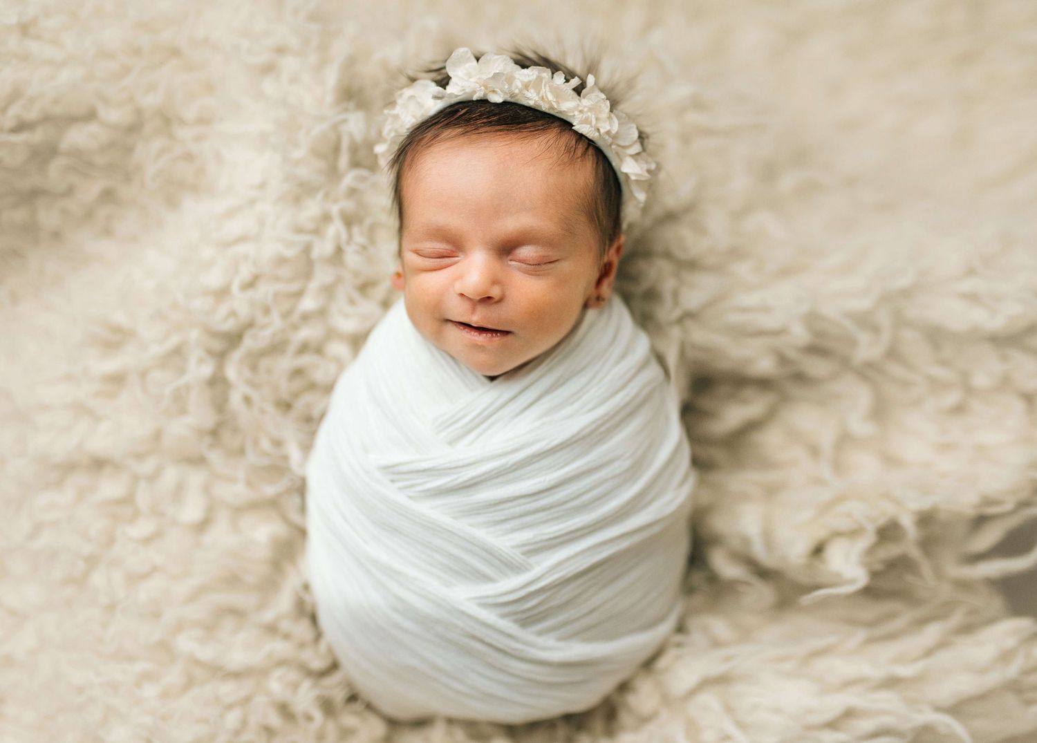 Sleeping newborn baby swaddled in white wrap with floral headband on fluffy beige backdrop.