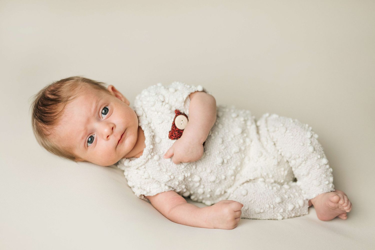 Adorable newborn baby in white textured romper lying on cream background with bright blue eyes.