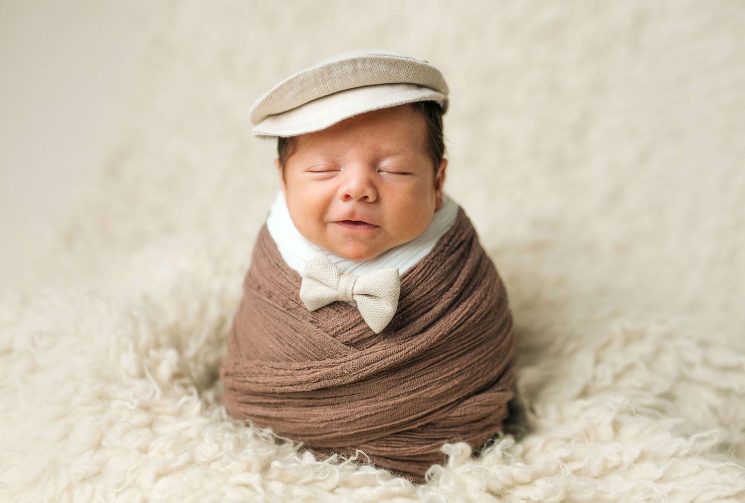 Newborn baby swaddled in brown wrap wearing a bow tie and flat cap, sleeping peacefully on a fluffy white rug.