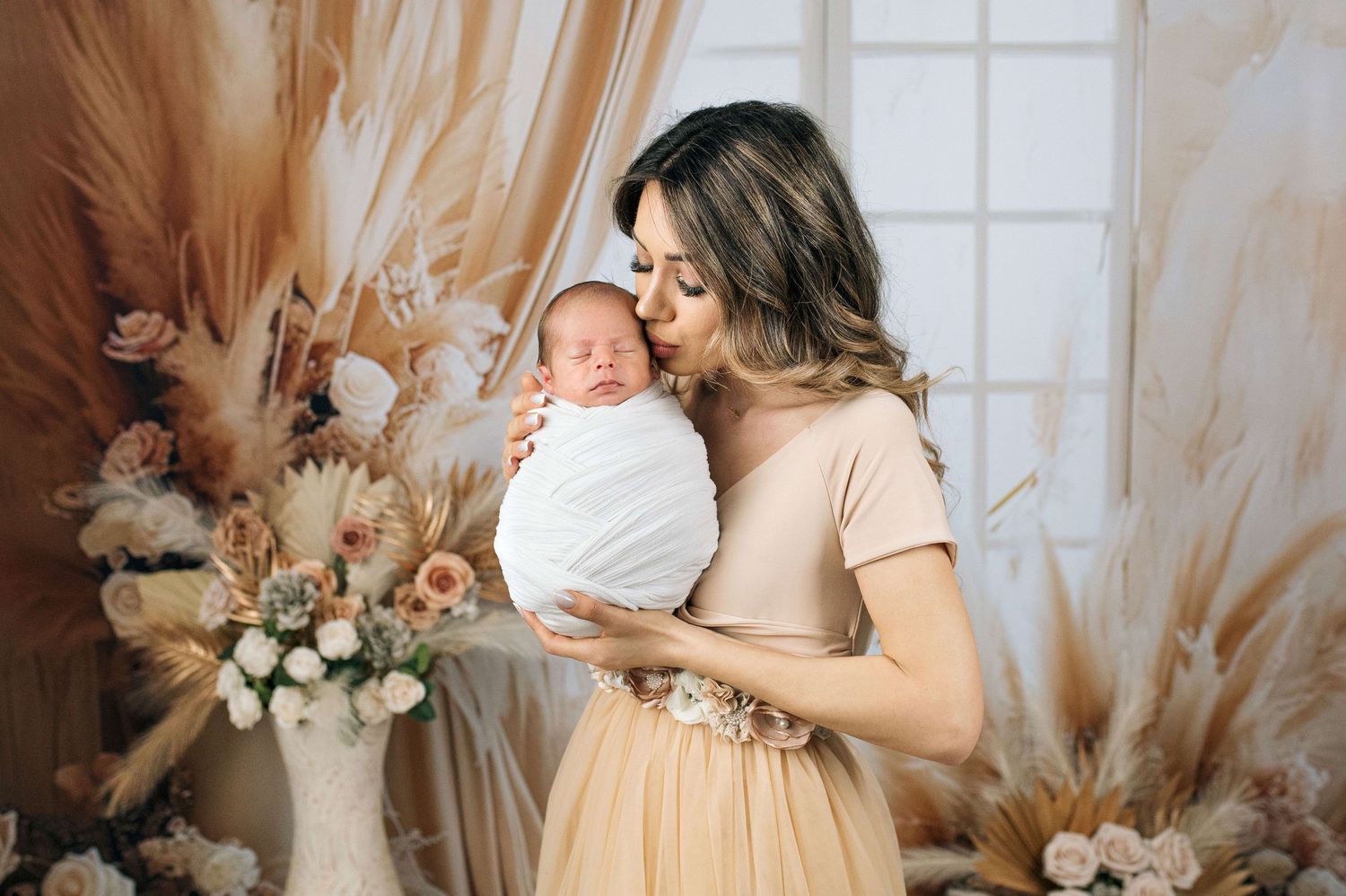 A mother in a beige dress tenderly kisses her swaddled newborn baby surrounded by boho floral arrangements.