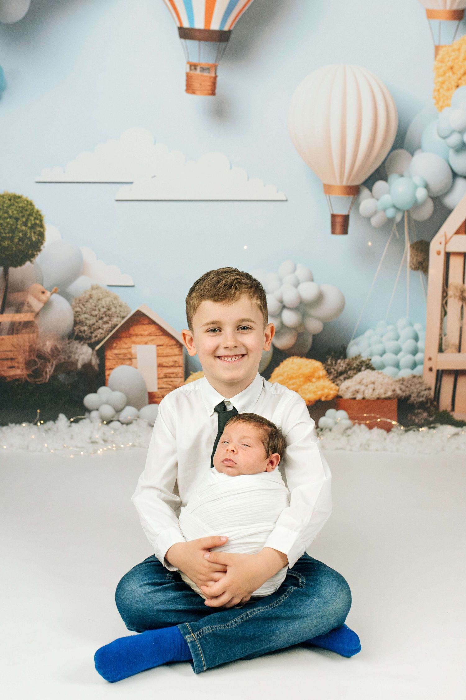Boy in white shirt holds swaddled newborn sibling, sitting cross-legged before hot air balloon nursery backdrop.