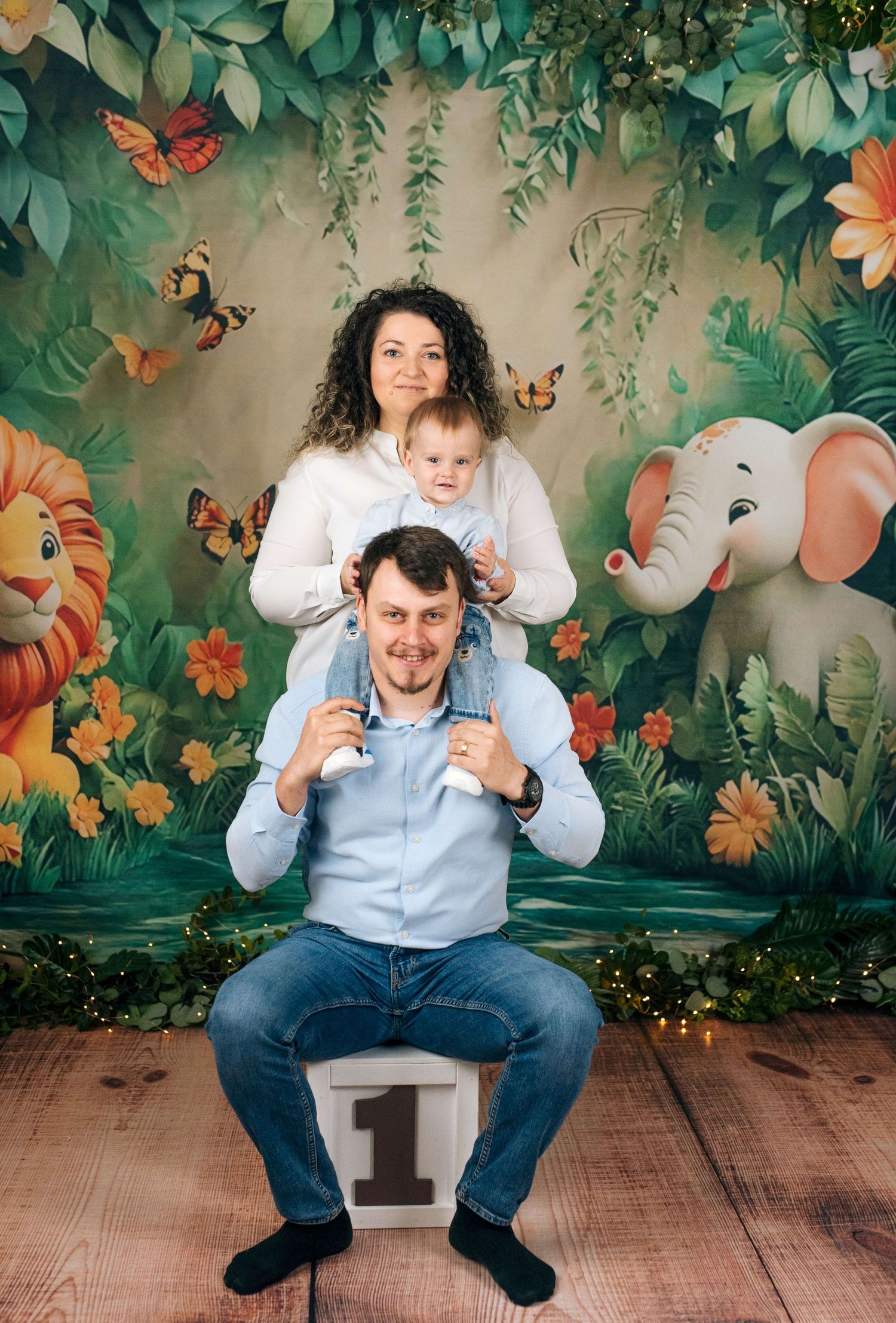 Family photo with parents and baby on jungle safari backdrop with lion, elephant, and butterflies for first birthday.