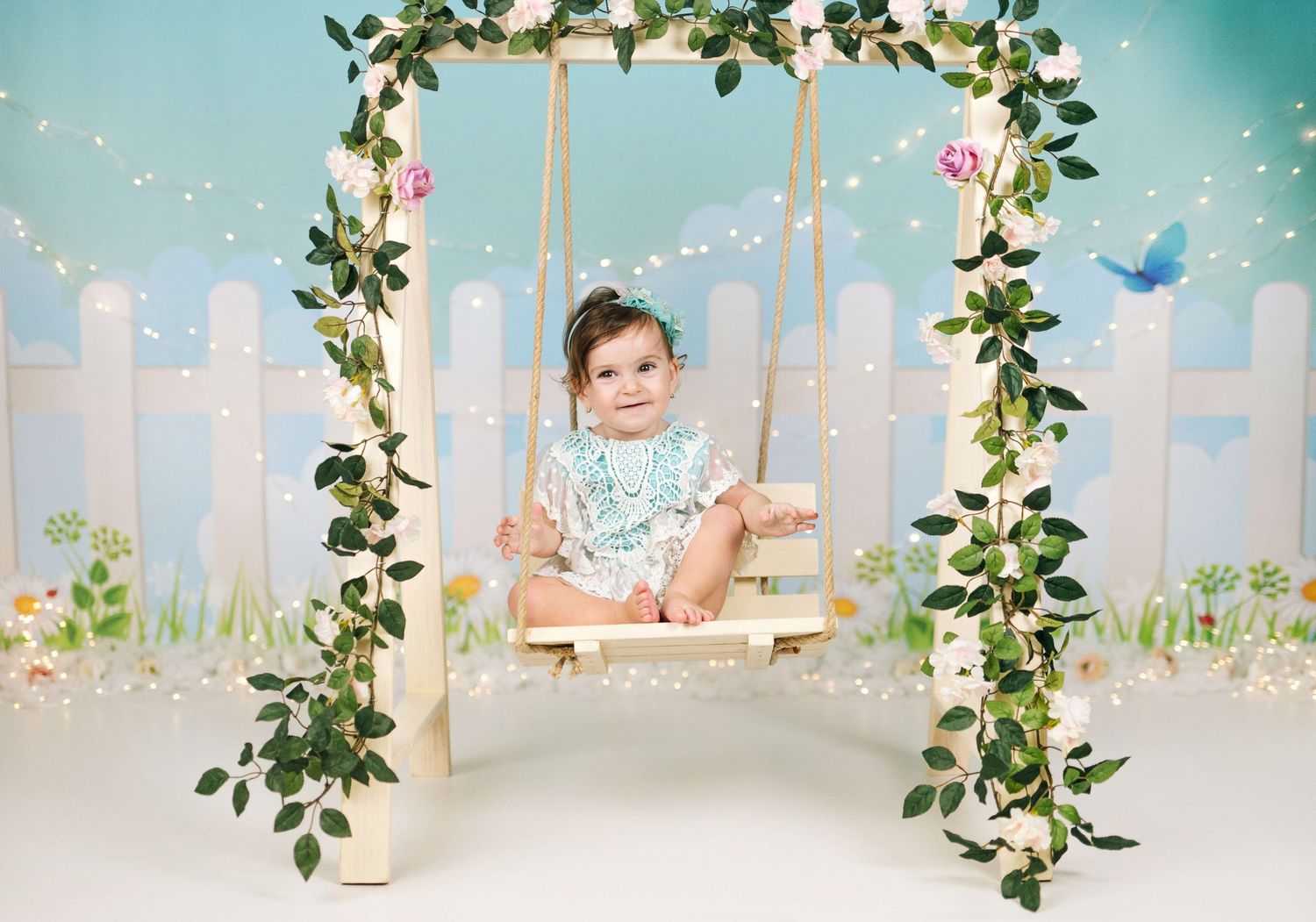 Baby girl in floral outfit sits on a decorated swing with greenery and fairy lights against a pastel garden backdrop.