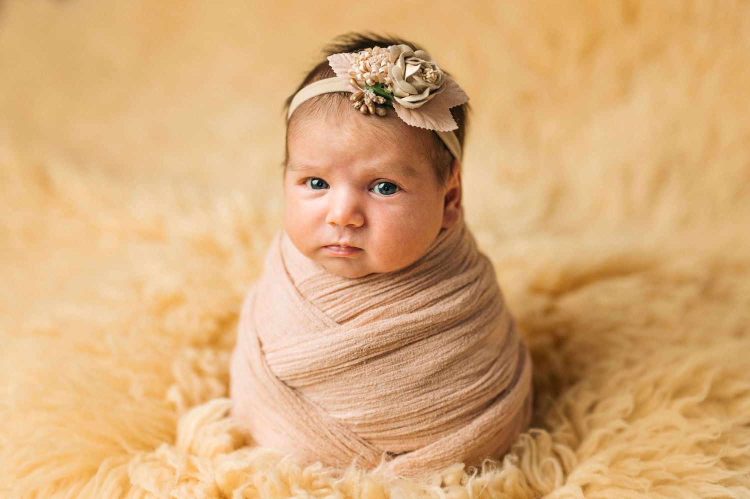 Newborn baby wrapped in peach swaddle with floral headband on fluffy yellow fur backdrop.