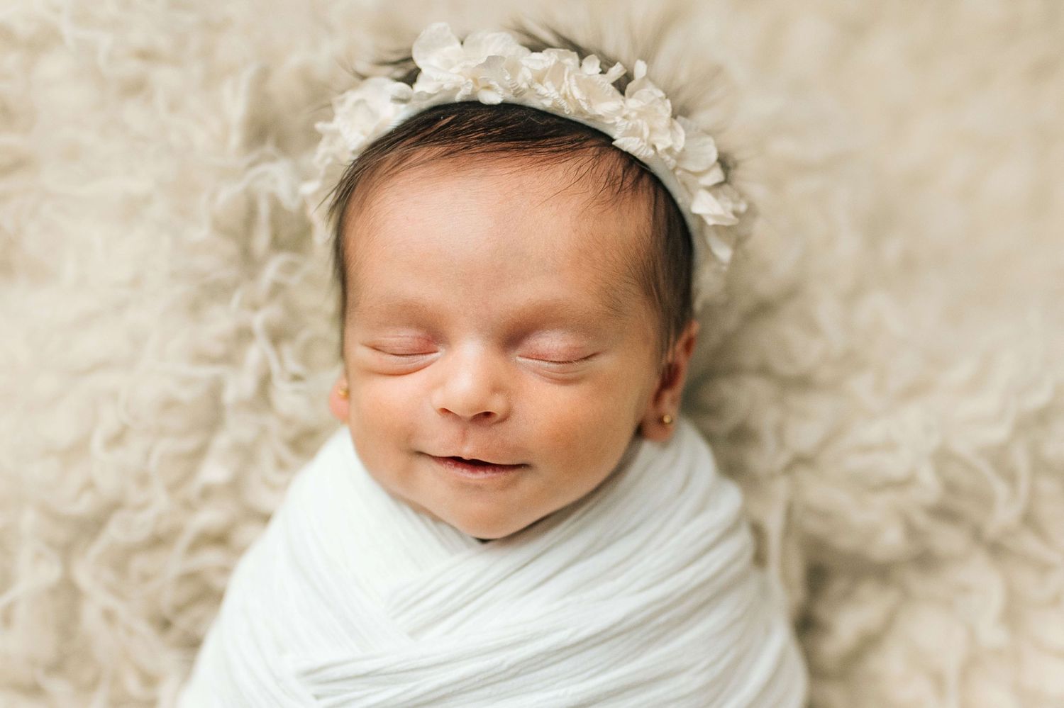 Sleeping newborn baby swaddled in white wrap wearing a delicate floral headband on a fluffy beige backdrop.