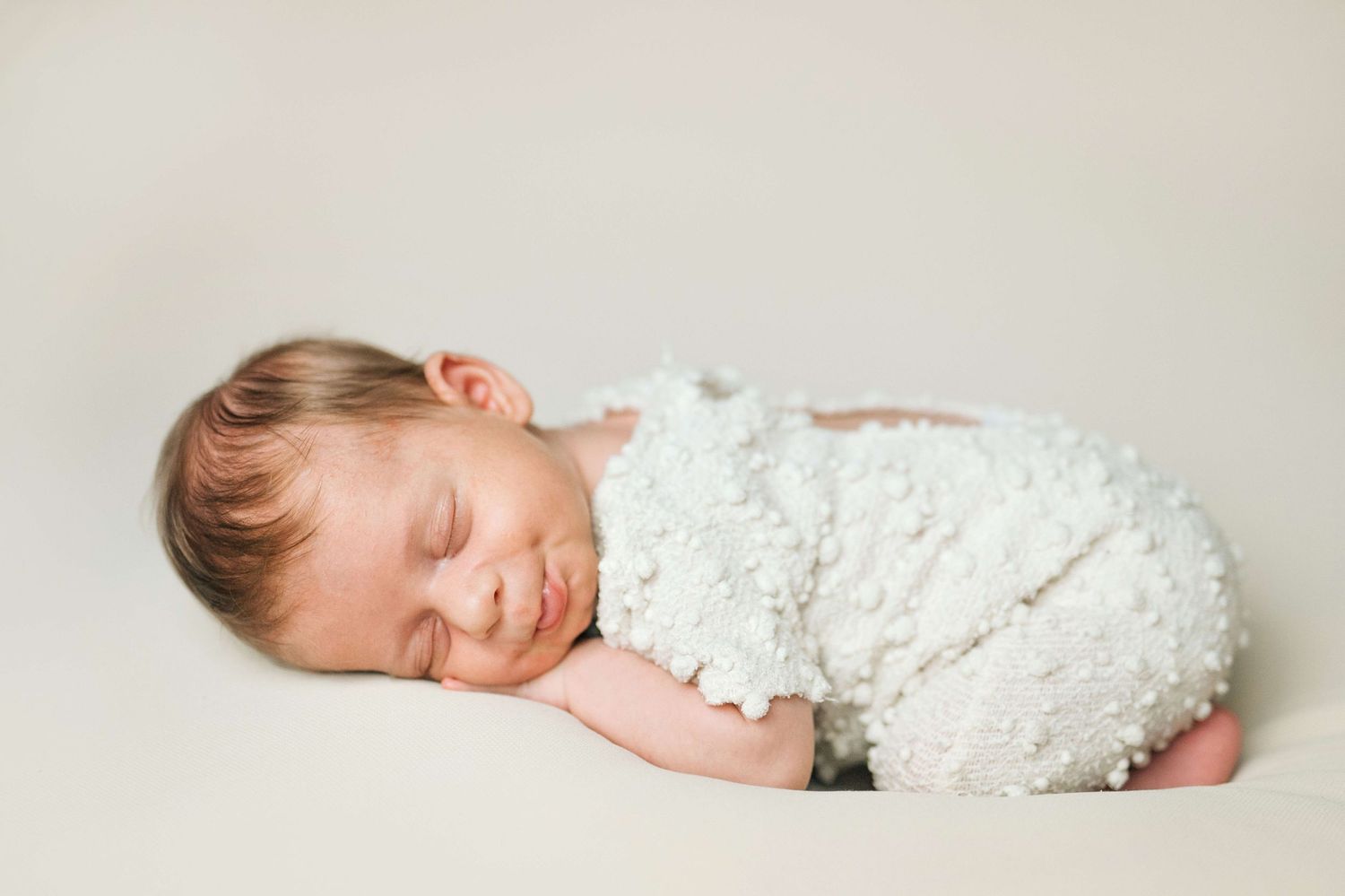 Peaceful newborn baby sleeping wrapped in a fluffy white blanket on a soft cream background.