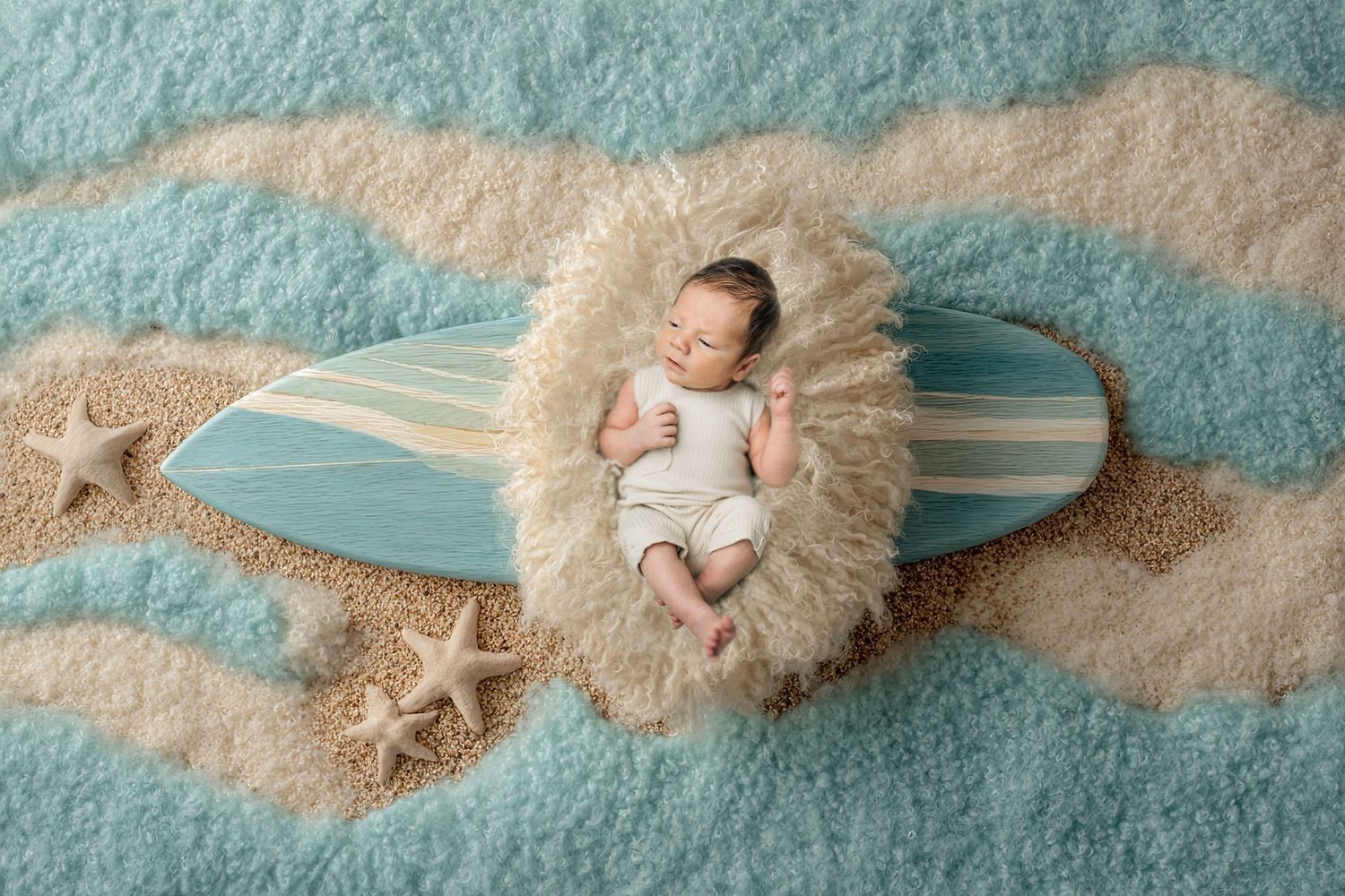 Newborn baby sleeping on a fluffy fur wrap atop a miniature surfboard, surrounded by starfish and sandy beach-themed backdrop.