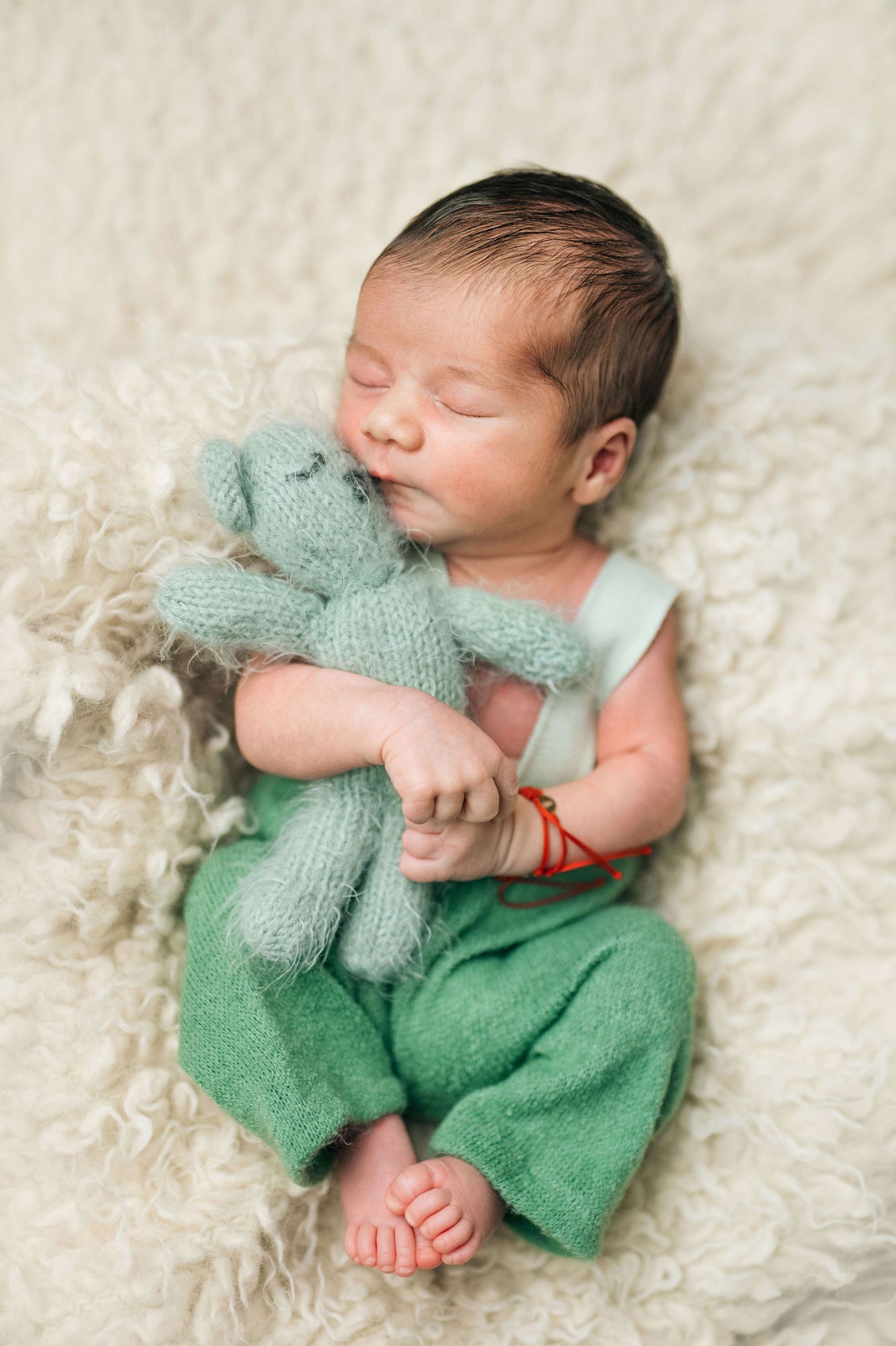 Sleeping newborn baby in green outfit cuddling a knitted teal teddy bear on a fluffy cream blanket.