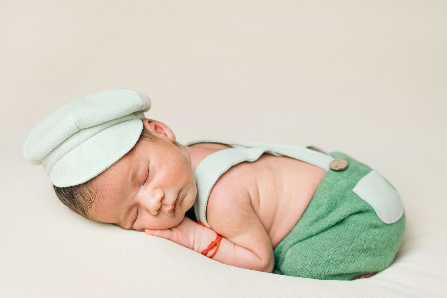 Newborn baby sleeping peacefully in green overalls and mint cap outfit on a soft beige background.