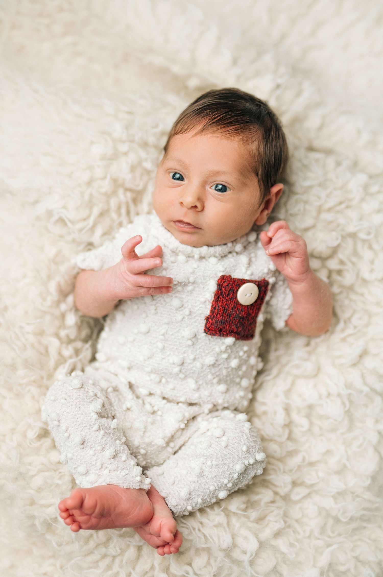 Newborn baby in white knit romper with red pocket lies on fluffy white blanket, looking at camera.