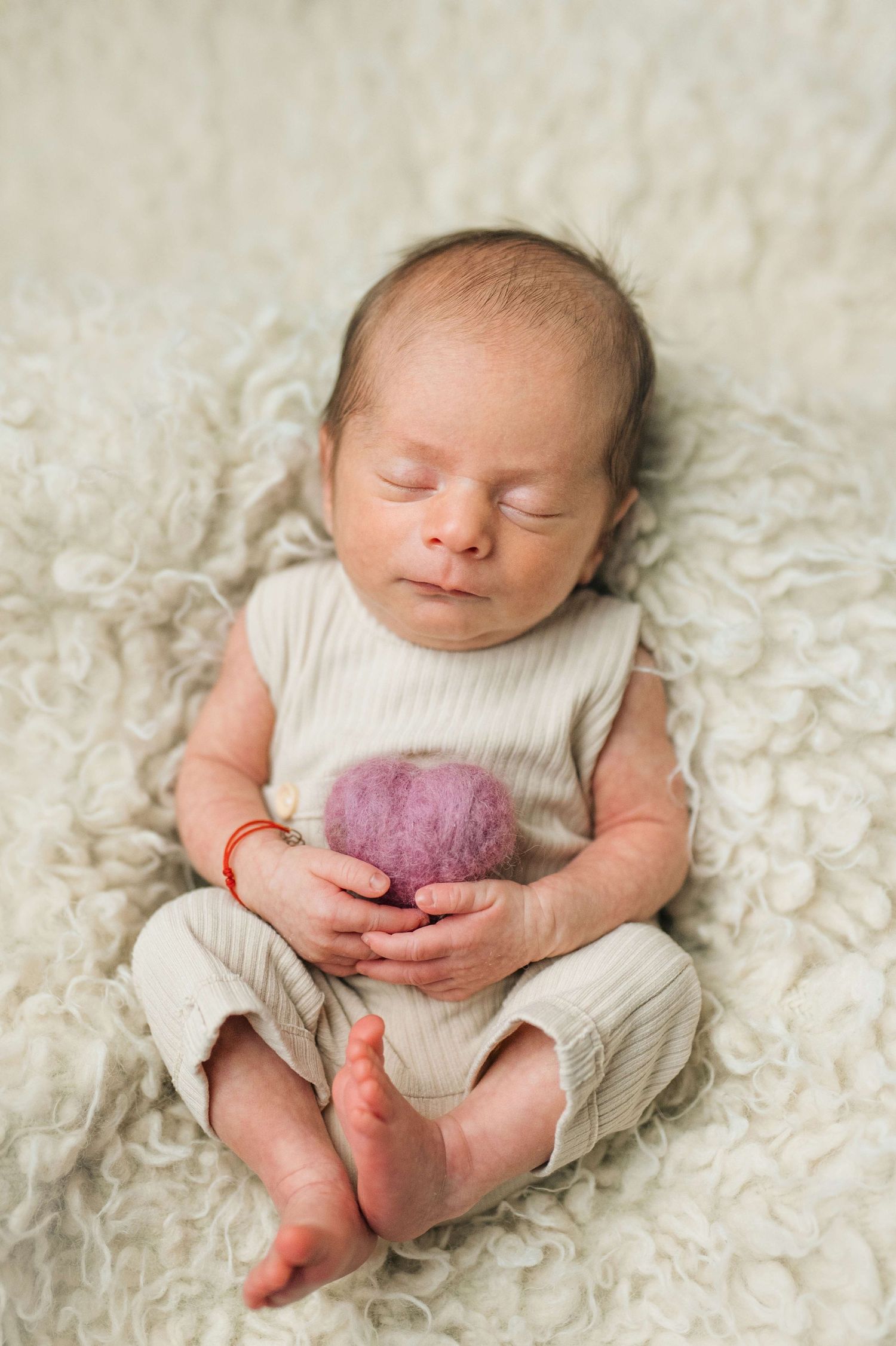 Sleeping newborn baby in cream ribbed outfit holding a small purple felt heart on a fluffy white blanket.