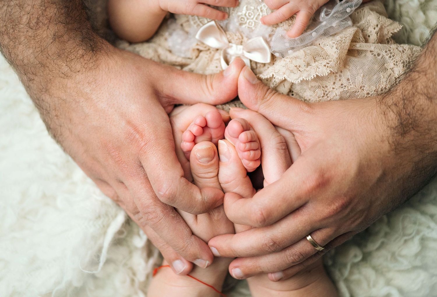 Parents gently cradling newborn baby feet in their hands, with tiny lace bow detail on soft white blanket.