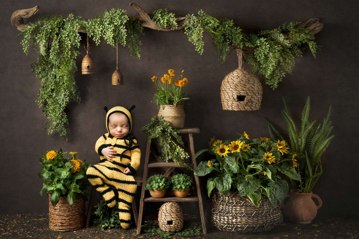 Baby in bee costume sits among sunflowers and potted plants with hanging beehives in a charming garden-themed photo.