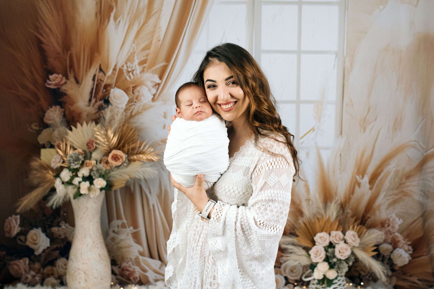 A smiling mother in a white lace dress holds her swaddled newborn baby surrounded by boho floral decor.