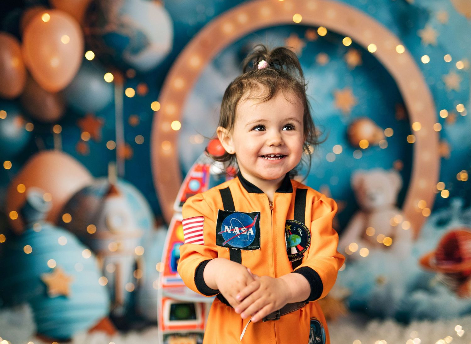 A smiling toddler girl wearing an orange NASA astronaut suit poses in front of a colorful space-themed backdrop with balloons.