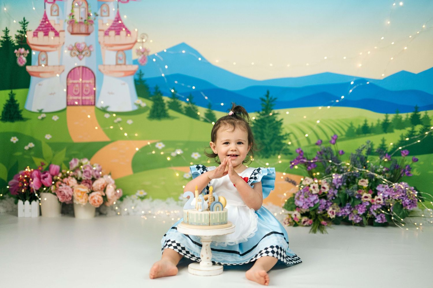 Baby girl in blue dress smiles at first birthday cake smash with fairy tale castle backdrop and flowers.