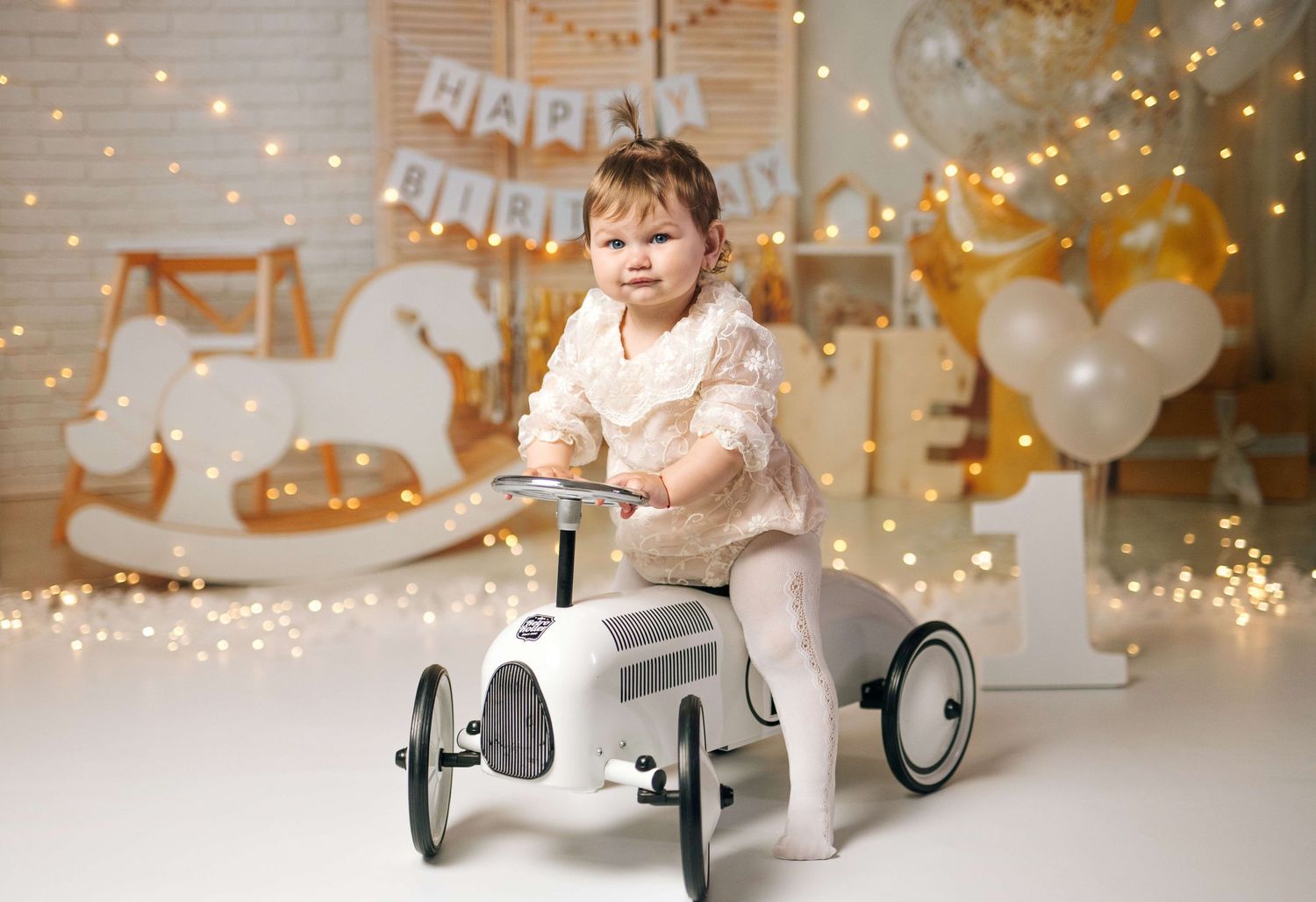 Toddler girl sitting on a vintage toy car at a Happy Birthday themed photo shoot with balloons and fairy lights.