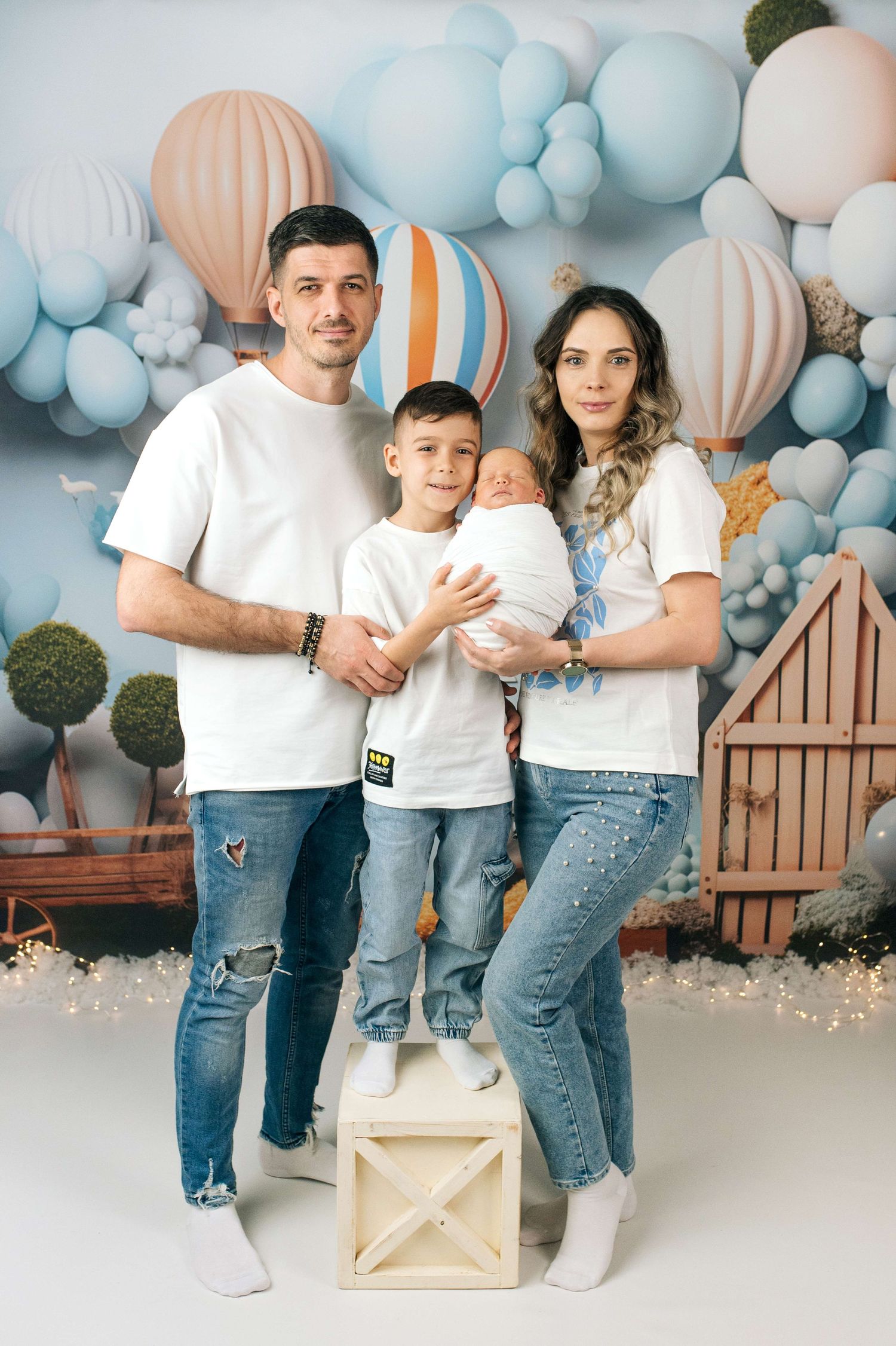 Family of four in white tees and jeans pose before a hot air balloon backdrop with a newborn baby.