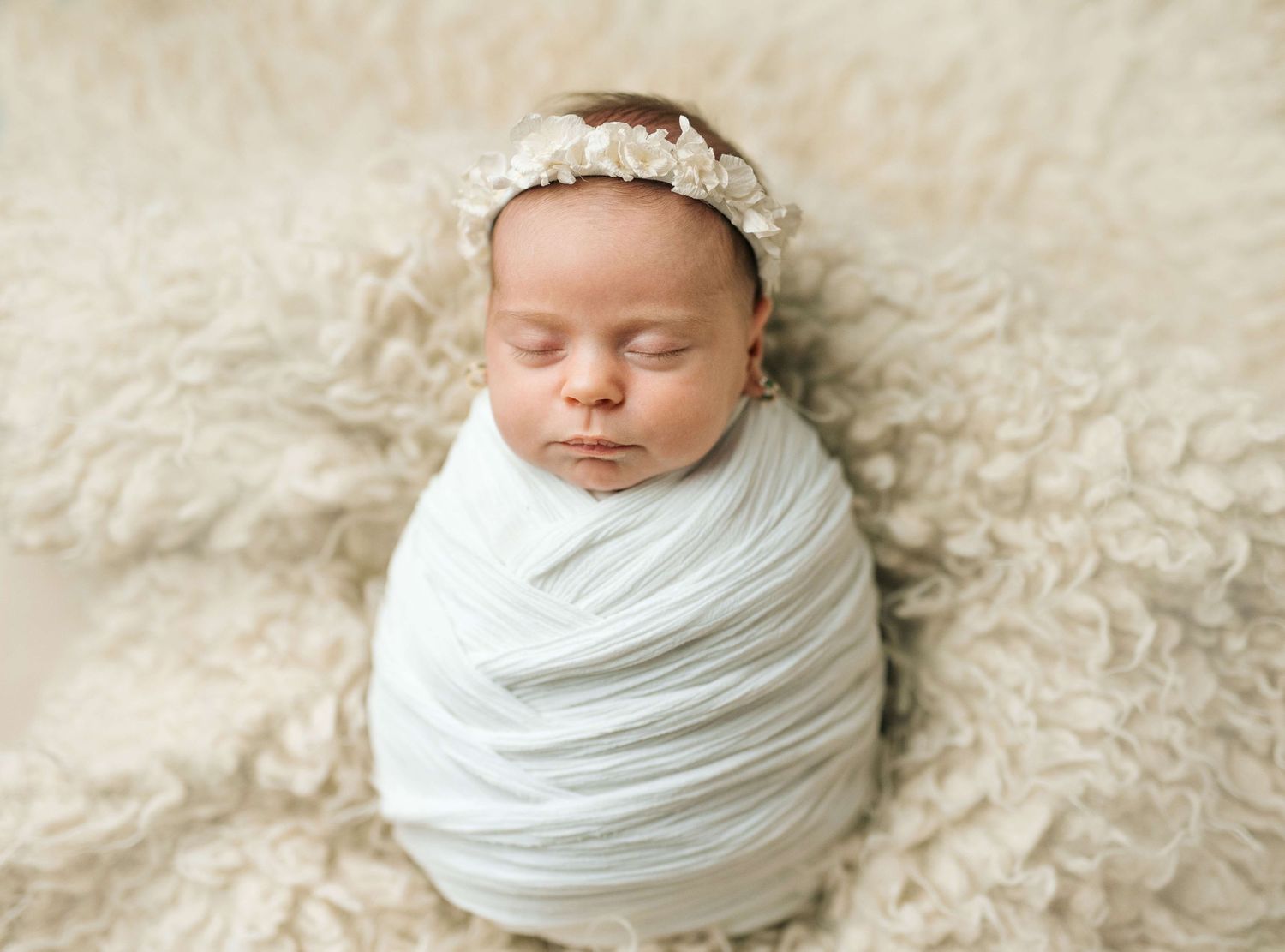 Newborn baby swaddled in white wrap with floral headband, sleeping peacefully on fluffy cream blanket.