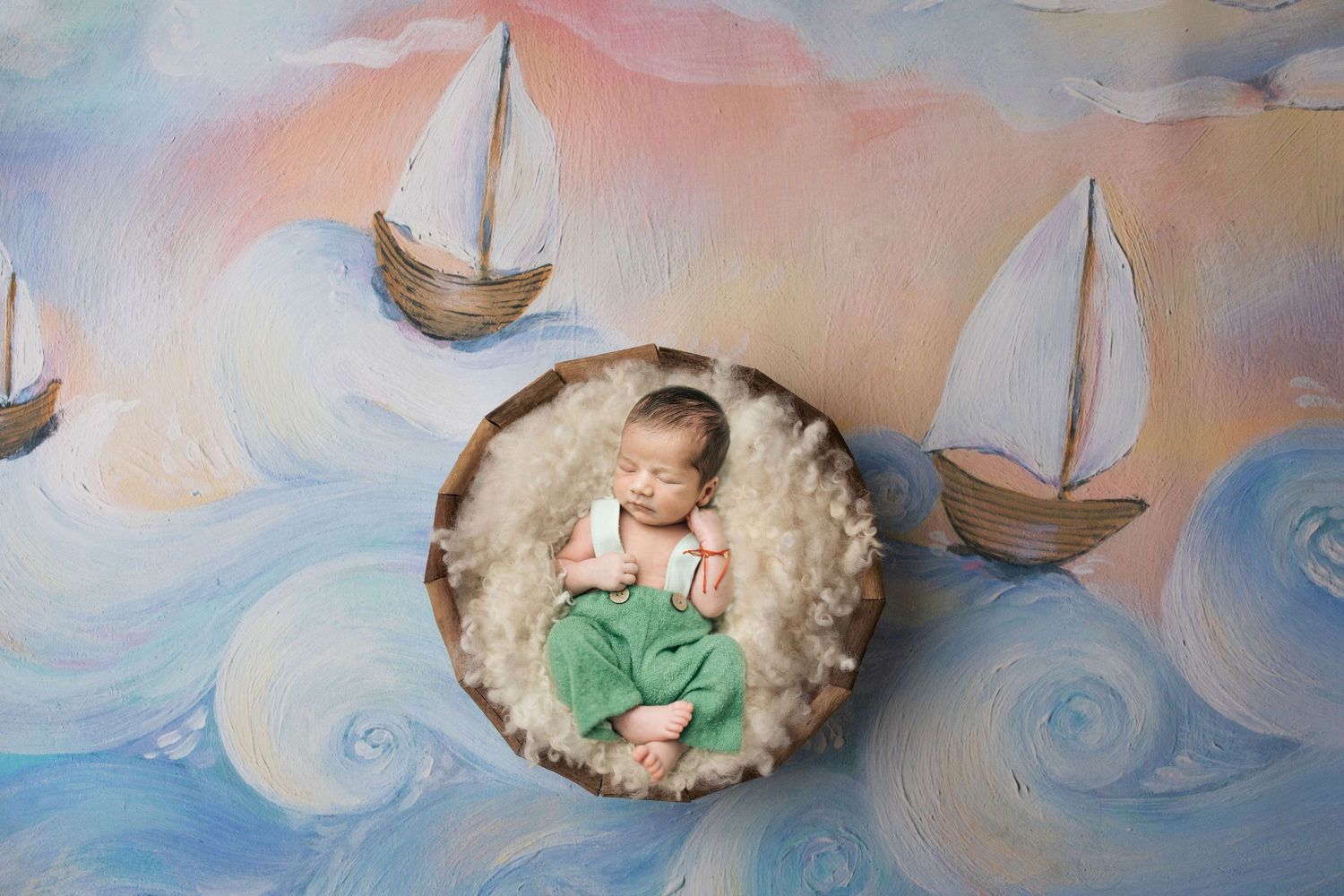 Newborn baby sleeping in a wooden bowl on fluffy white fur, surrounded by a painted sailboat ocean backdrop.