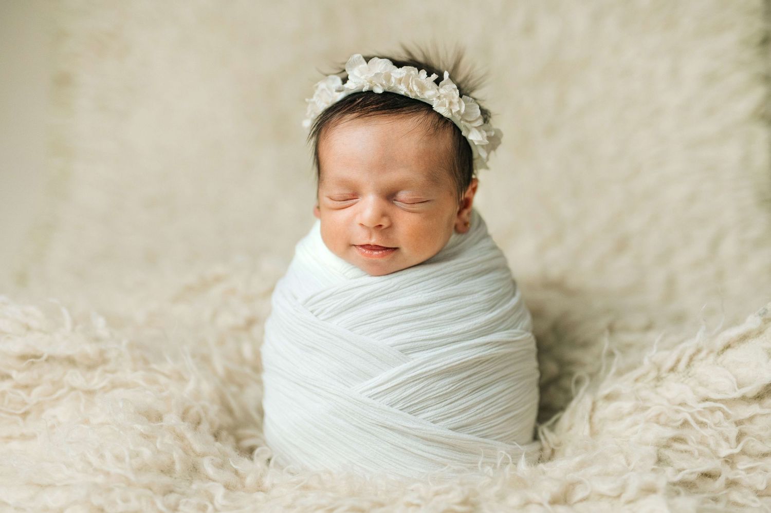 Sleeping newborn baby swaddled in white wrap wearing floral crown on fluffy cream backdrop.