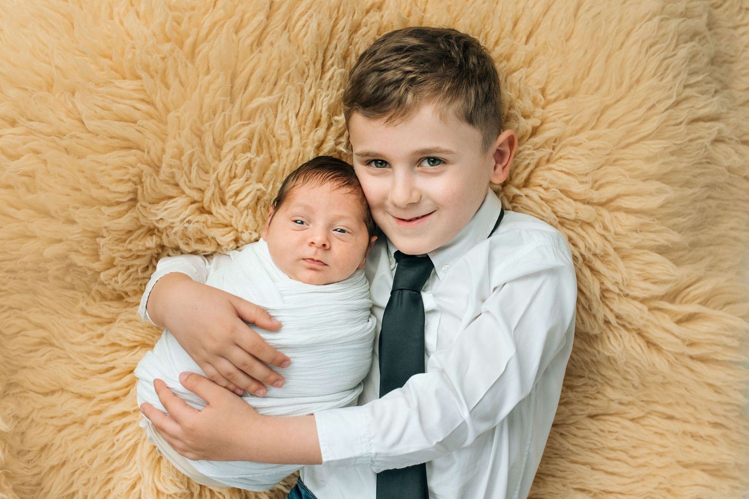Older boy in white shirt and tie tenderly holds swaddled newborn baby on fluffy beige background.