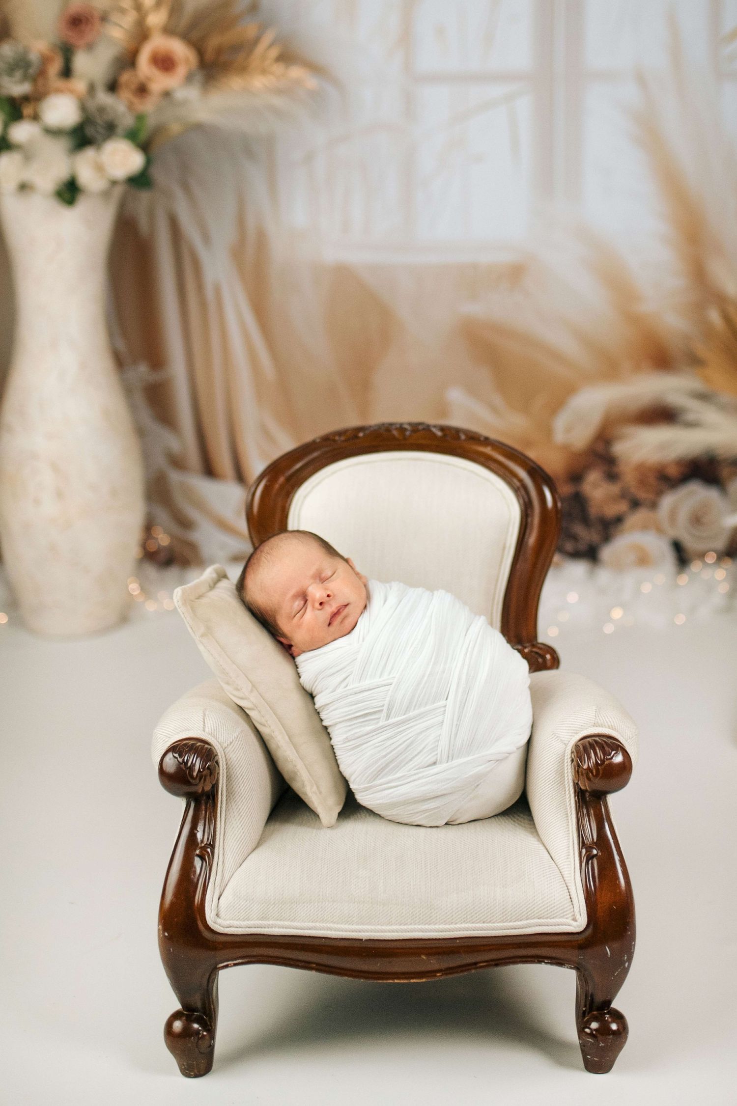Sleeping newborn baby swaddled in white wrap resting on a miniature cream armchair in a soft boho studio setting.