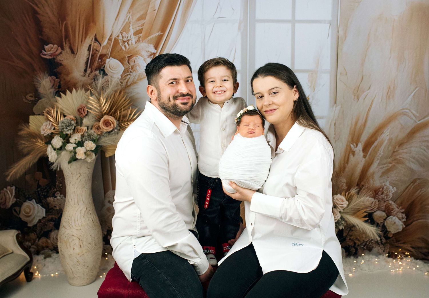 Happy family portrait with parents and two children, including a newborn baby, posed before a floral backdrop.