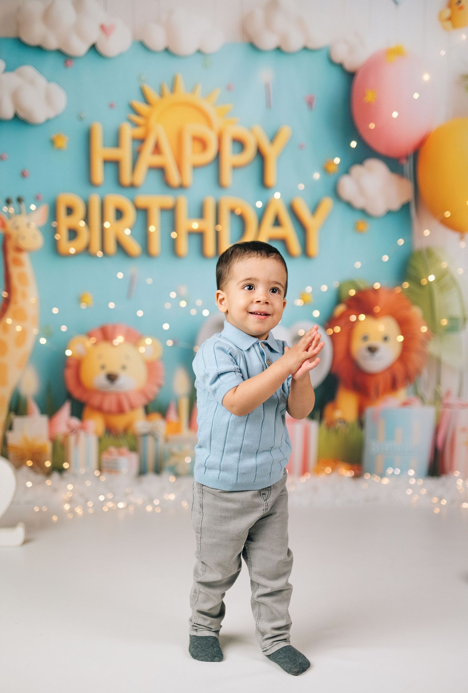 A smiling toddler boy claps hands at a colorful Happy Birthday backdrop with lions, giraffes, and balloons.
