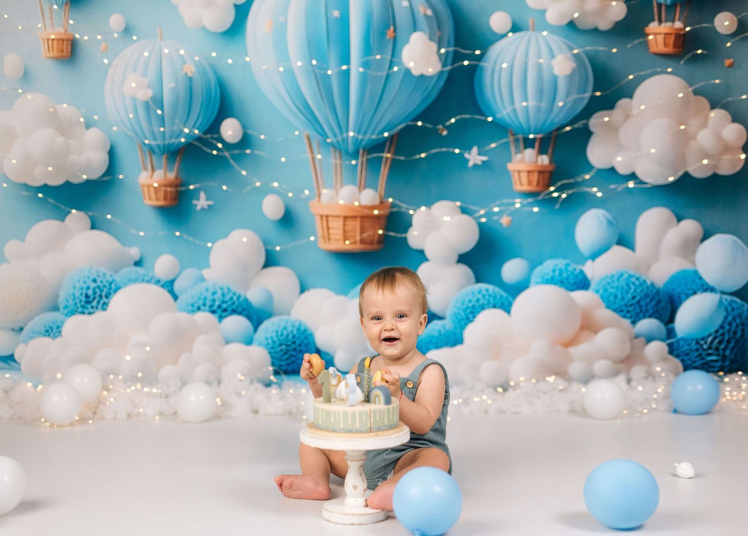 Baby smiling at cake smash session with blue hot air balloon backdrop, clouds, and balloons for first birthday celebration.