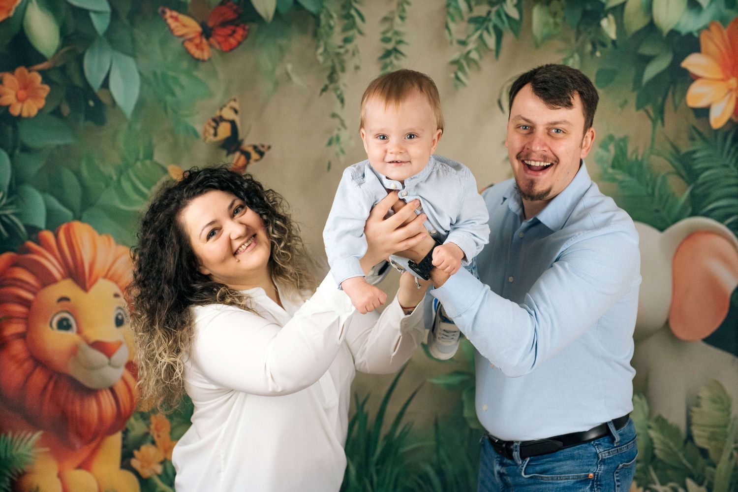 Happy family with curly-haired mom, smiling dad, and baby boy posing in front of colorful jungle animal backdrop.