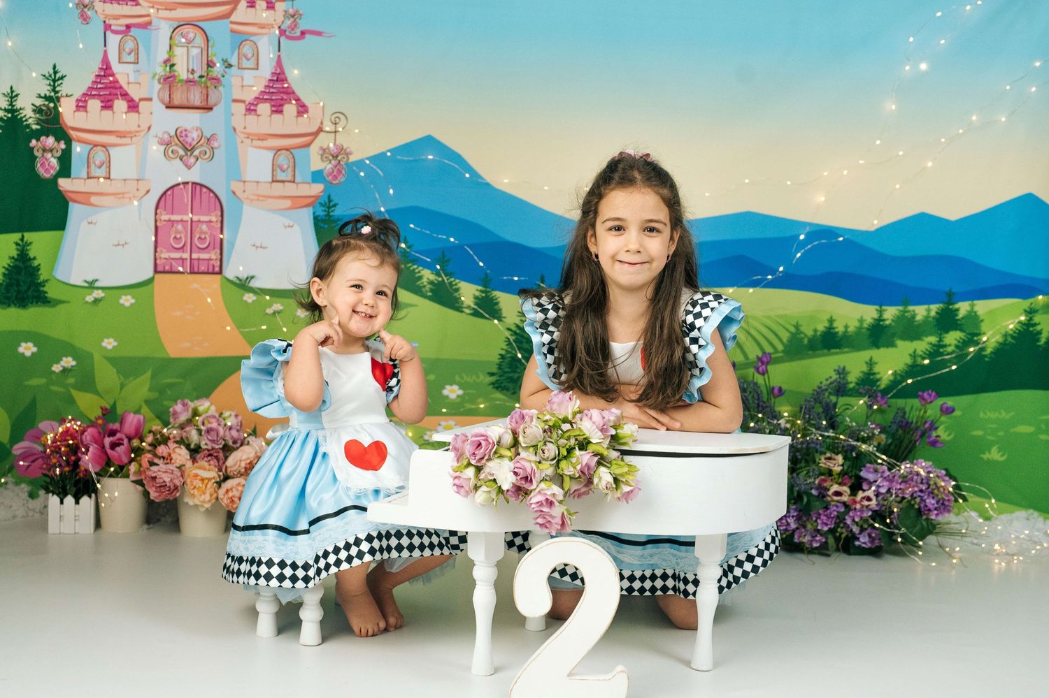 Two young girls in Alice in Wonderland dresses pose by a white piano with a fairy tale castle backdrop.