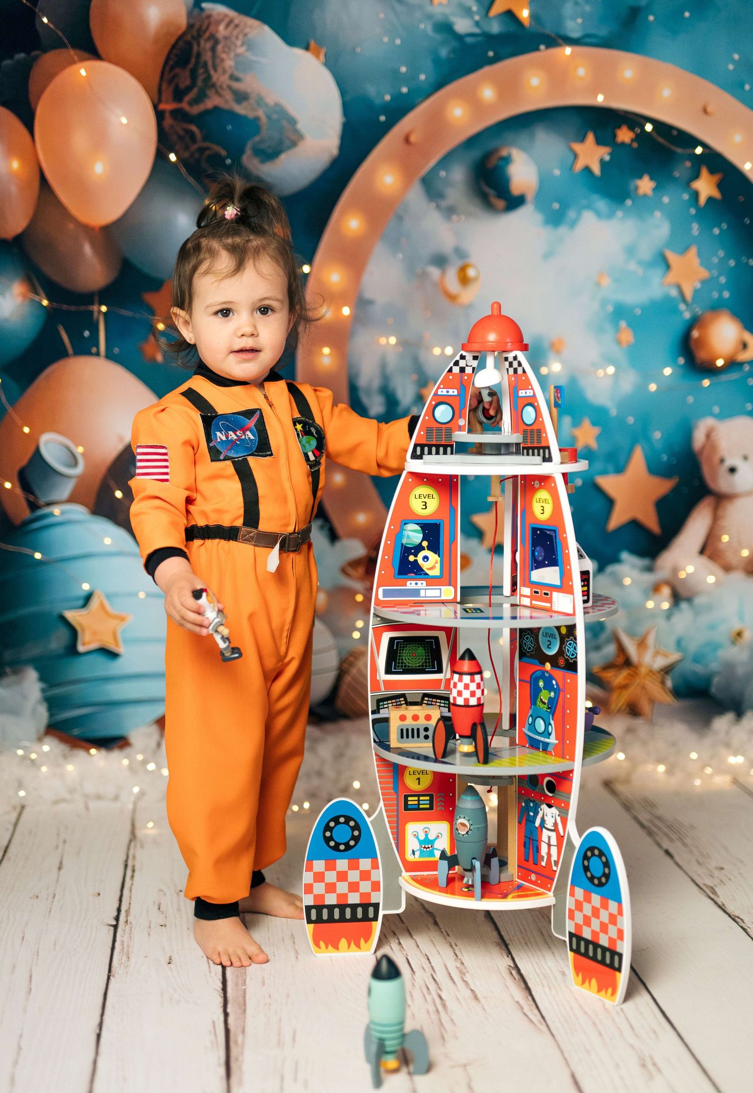 Toddler in NASA astronaut costume poses with colorful rocket toy playset against space-themed backdrop with balloons and stars.