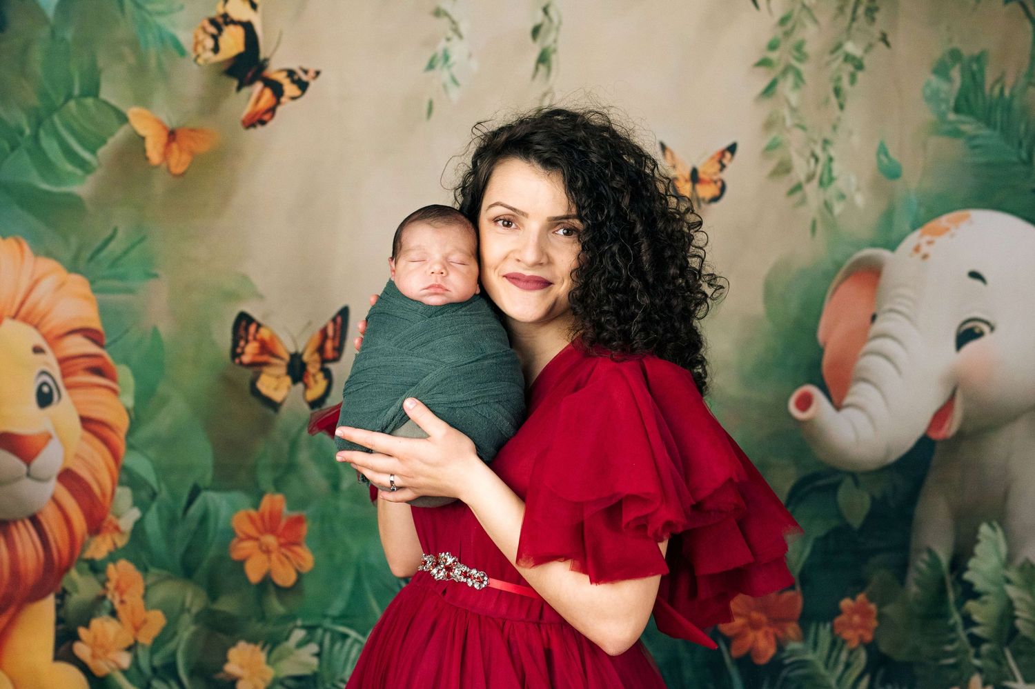 A woman in a red dress holds a swaddled newborn baby against a colorful jungle animal backdrop.