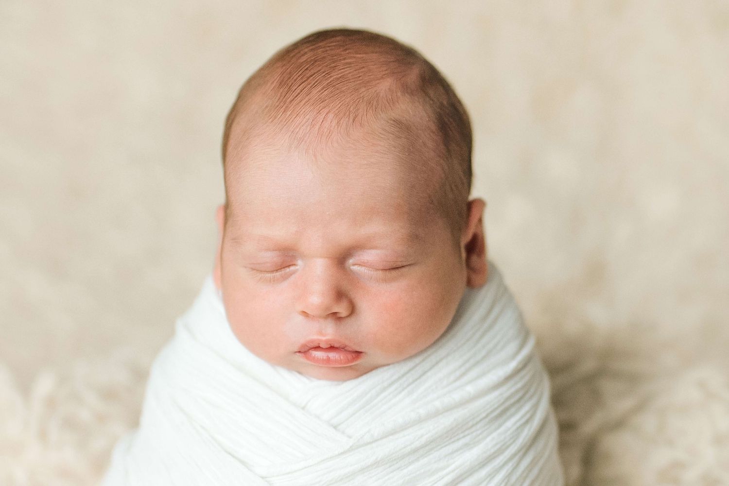 Peaceful newborn baby swaddled in white wrap sleeping on cream background, professional newborn photography.