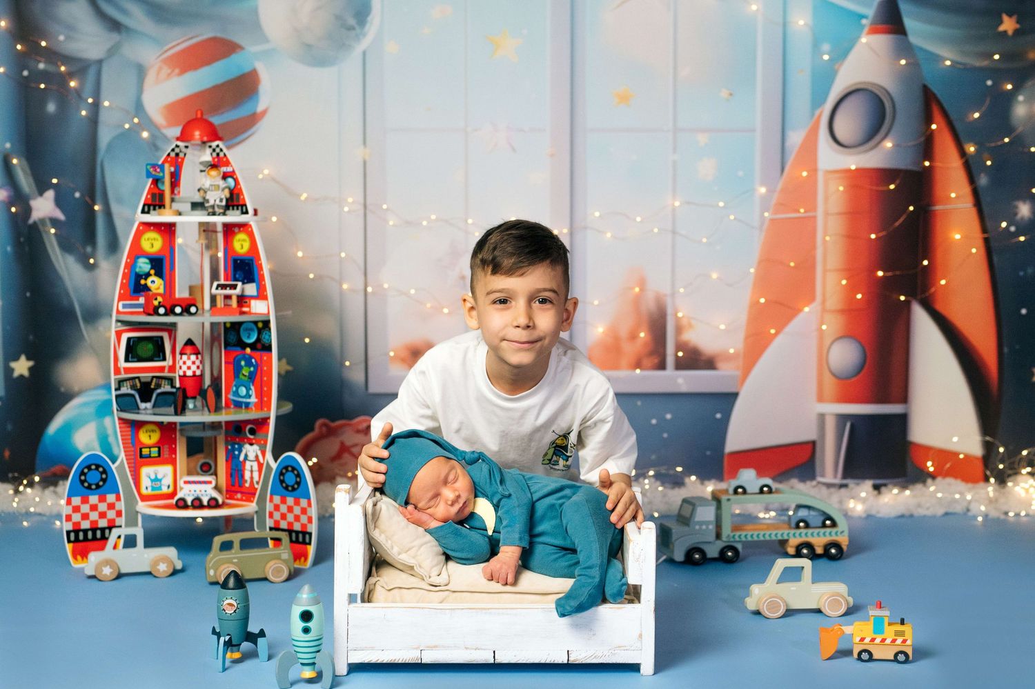 Older boy proudly holds sleeping newborn sibling on white bed surrounded by colorful rocket ship toys and space-themed backdrop.