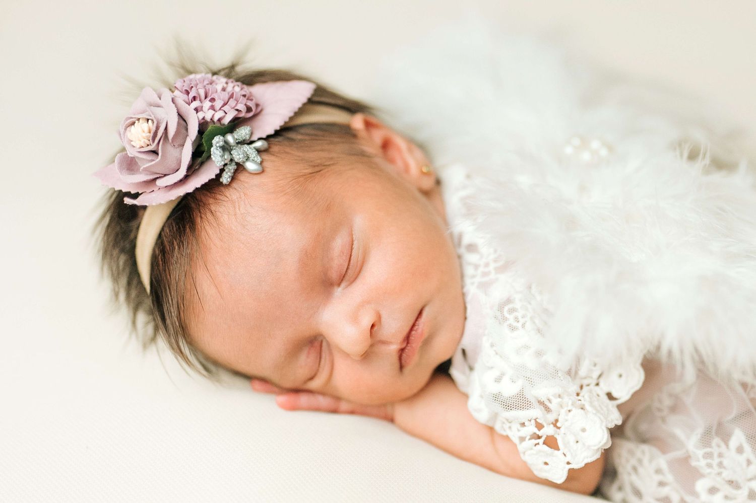 Sleeping newborn baby girl wearing a floral headband, wrapped in white lace and feathers.